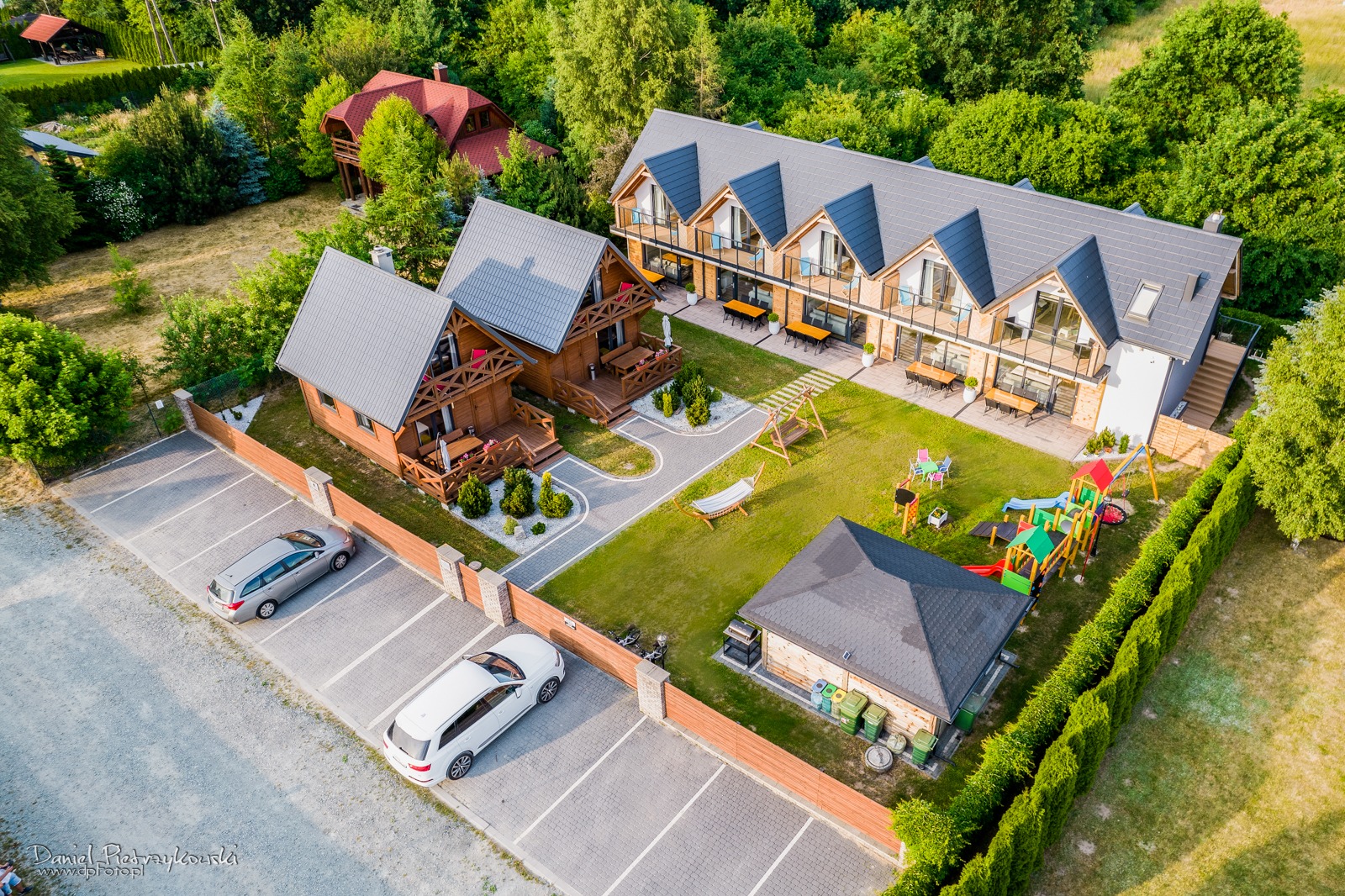 Widok z lotu ptaka na kompleks drewnianych domków z parkingiem, placem zabaw i miejscem do wypoczynku na trawie.