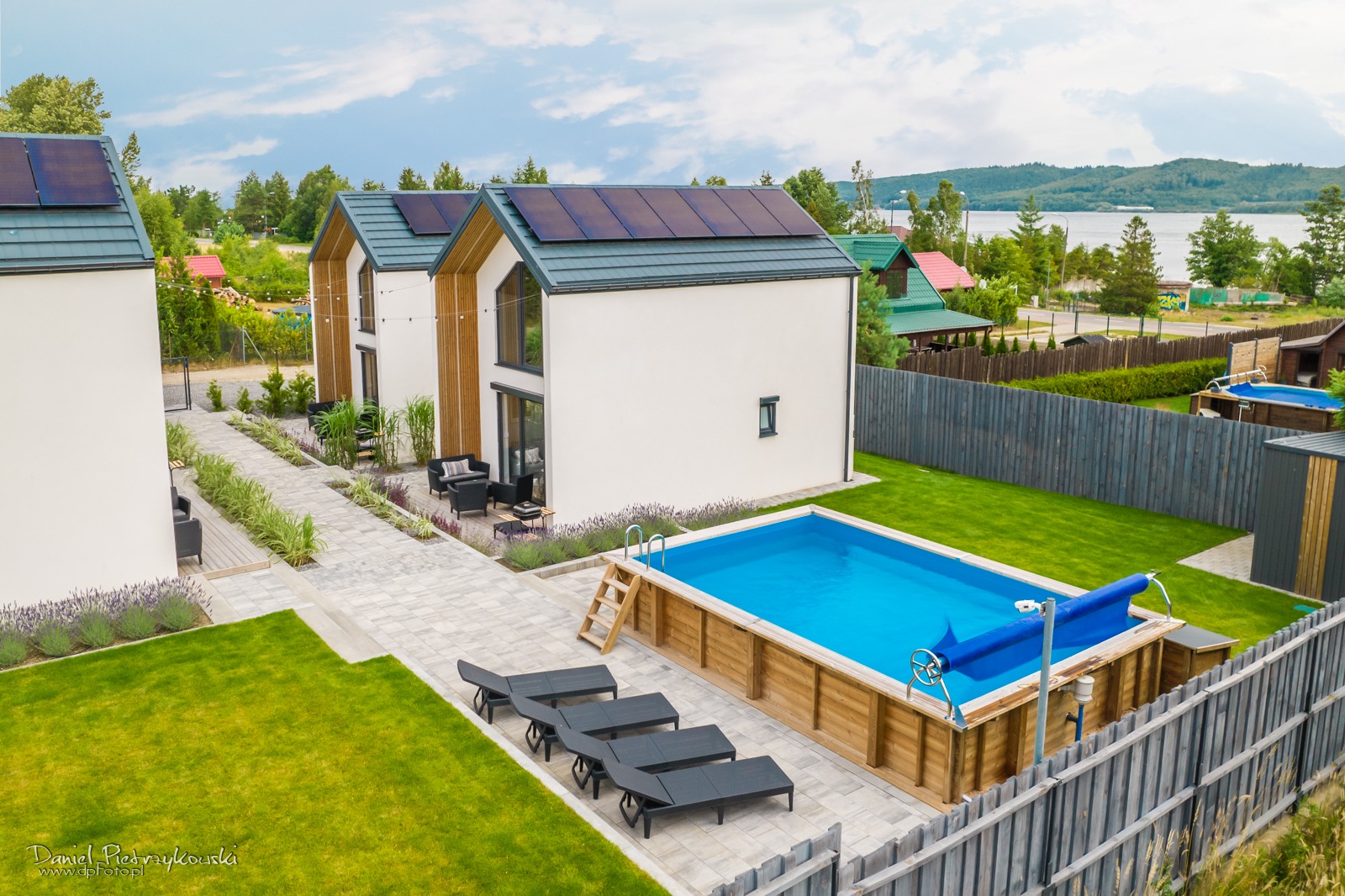 Nowoczesne domy jednorodzinne z panelami słonecznymi, basenem naziemnym, leżakami i zagospodarowanym terenem zielonym z widokiem na jezioro w tle.