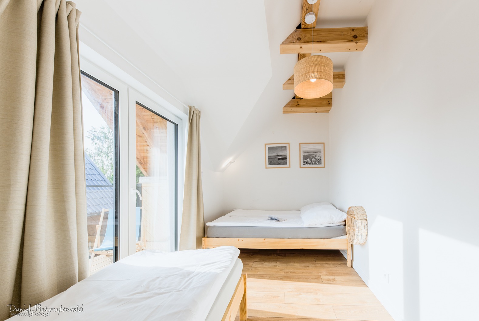 Jasne wnętrze pokoju z dwoma łóżkami, drewnianą podłogą, oknem z zasłonami i lampą wiszącą z rattanu