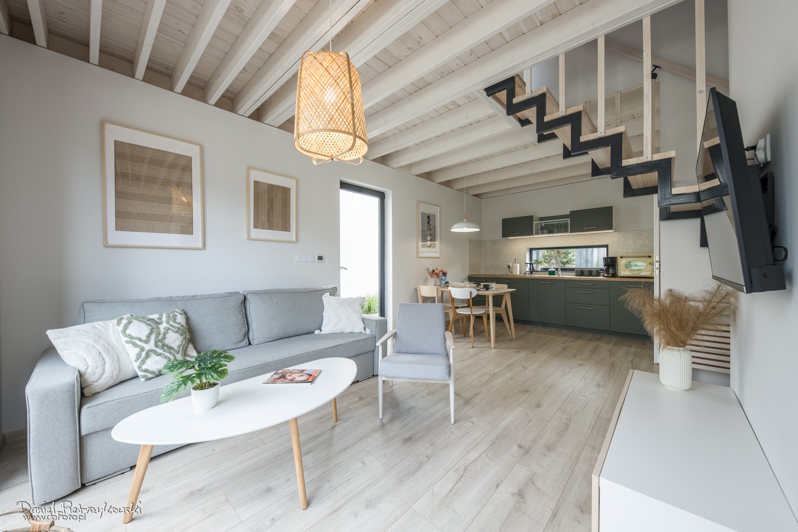 Nowoczesne wnętrze mieszkania z otwartą przestrzenią: szara sofa z poduszkami, biały stolik kawowy z rośliną, drewniane podłogi, kuchnia w odcieniach zieleni i schody z metalowymi elementami.