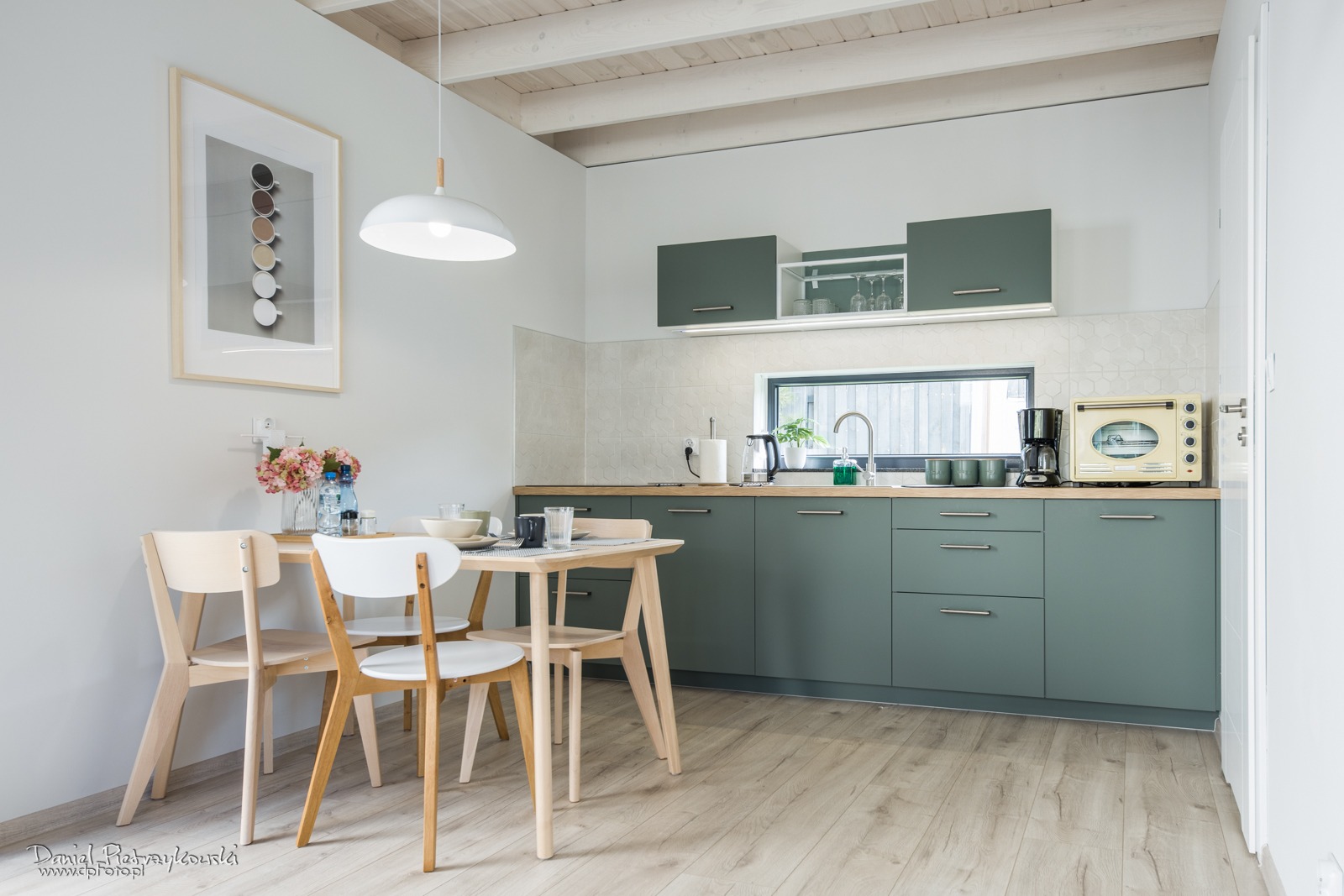 Jasna kuchnia z drewnianym stołem i krzesłami, zielone szafki, beżowe płytki, drewniana podłoga, minimalistyczny obraz na ścianie.