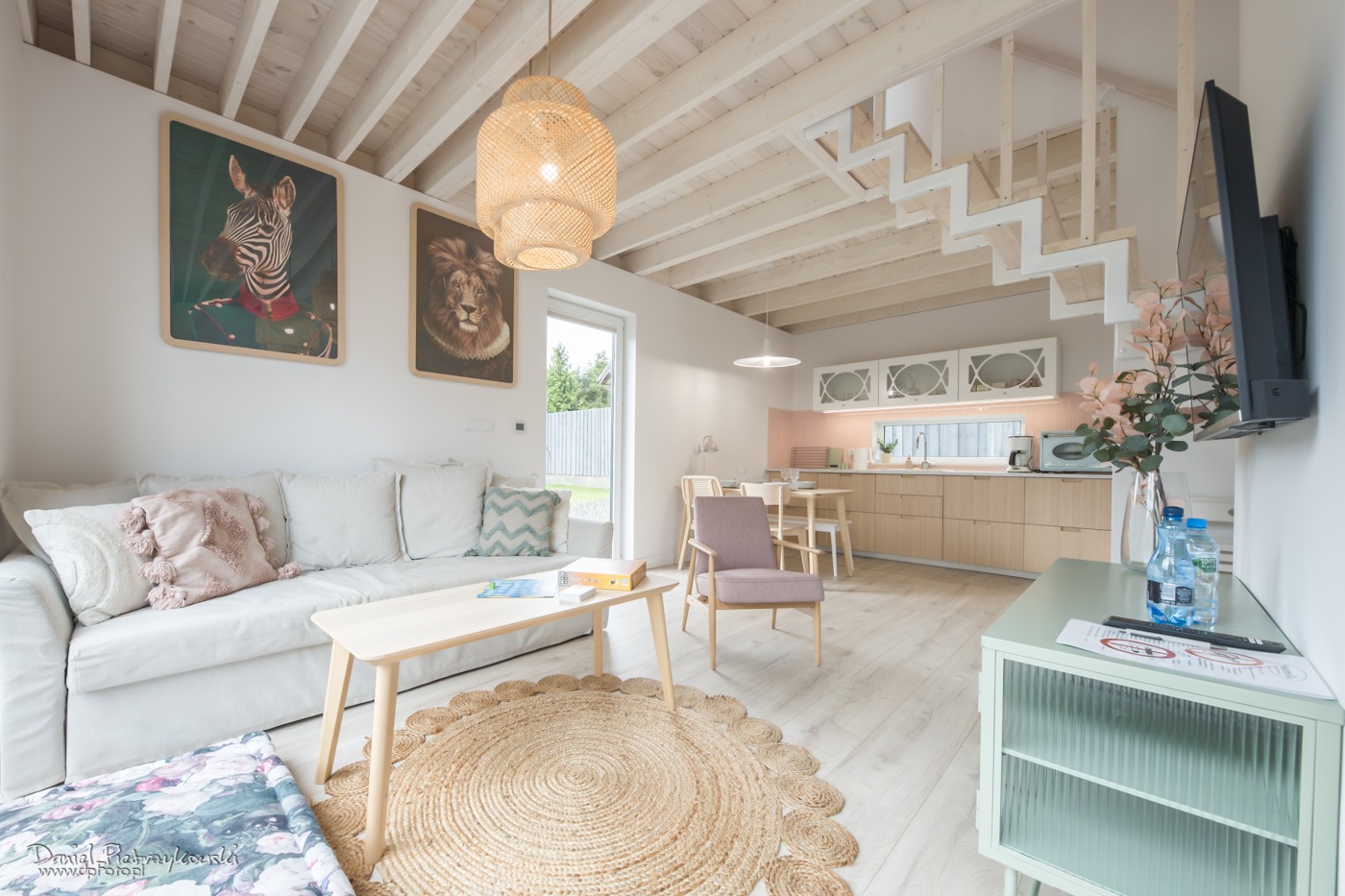 Jasne wnętrze domu z drewnianym wykończeniem, obejmujące salon z sofą, stolikiem kawowym na okrągłym dywanie, kuchnię z jadalnią w tle oraz schody bez poręczy prowadzące na górę; na ścianach wiszą...