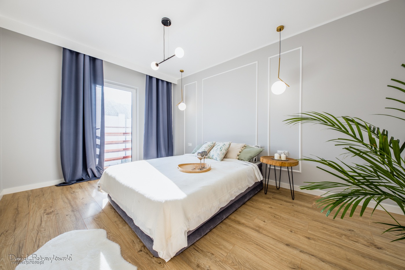 Jasna sypialnia w stylu minimalistycznym z drewnianą podłogą, szarymi ścianami z dekoracyjnymi listwami, łóżkiem z białą pościelą, stolikiem nocnym z drewna i lampami wiszącymi. Na łóżku taca...