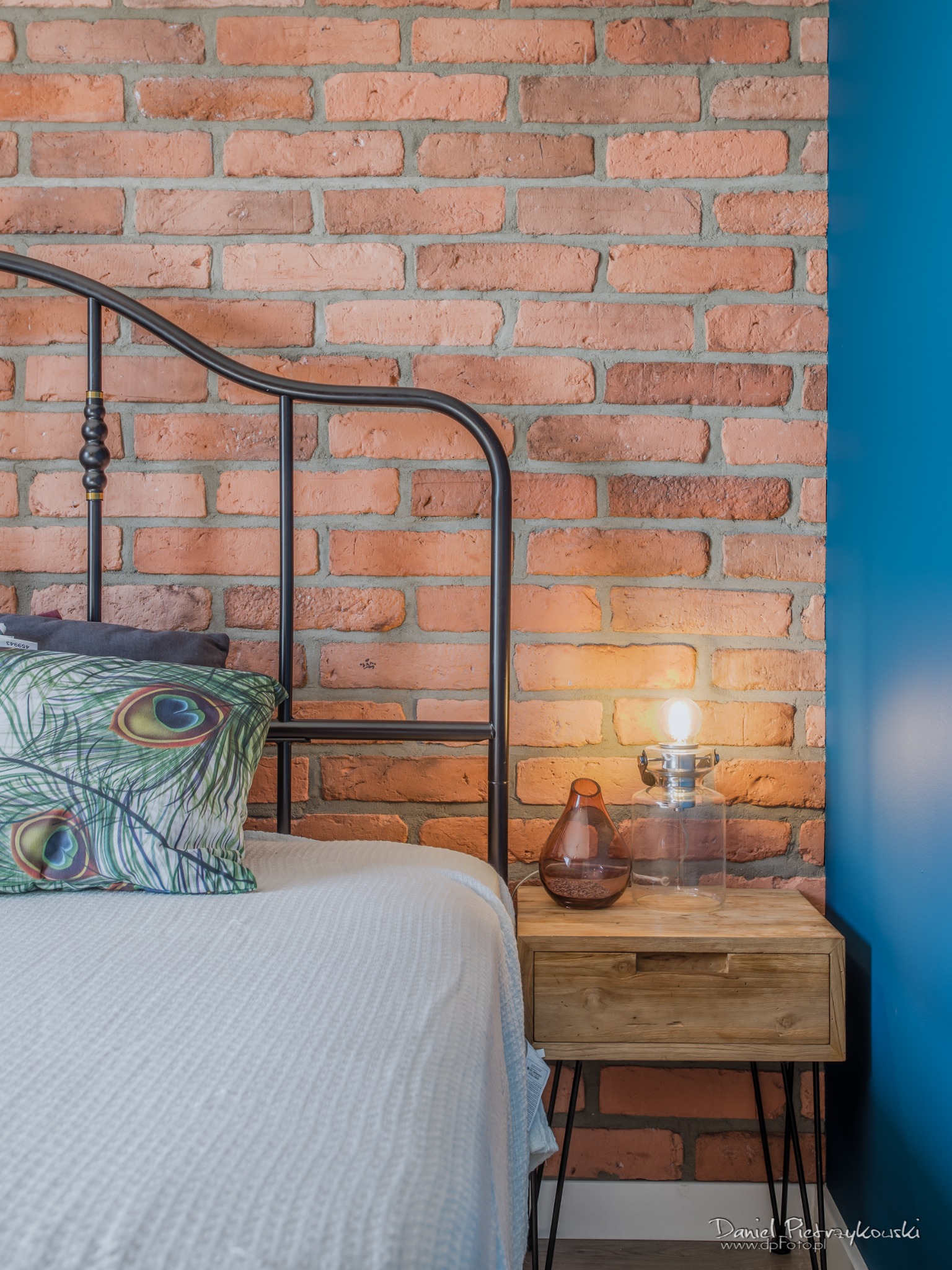 Sypialnia w stylu loft: metalowe łóżko z poduszką w pawie pióra, ceglana ściana, drewniana szafka nocna z lampką i dekoracyjnym wazonem, niebieska ściana w tle.