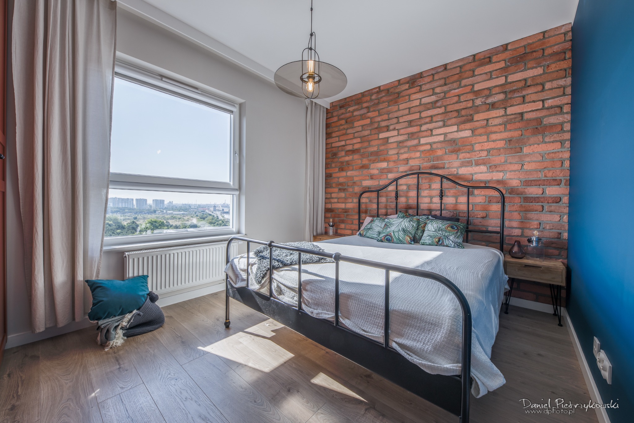 Sypialnia w stylu loft z łóżkiem z metalową ramą, ceglaną ścianą i widokiem na miasto za oknem.