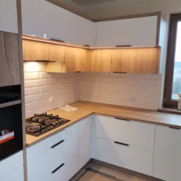 Nowoczesna kuchnia z białymi frontami i drewnianymi akcentami, białe płytki w stylu metro, płyta gazowa, oświetlenie LED pod szafkami górnymi, widoczne elementy montażowe na podłodze.