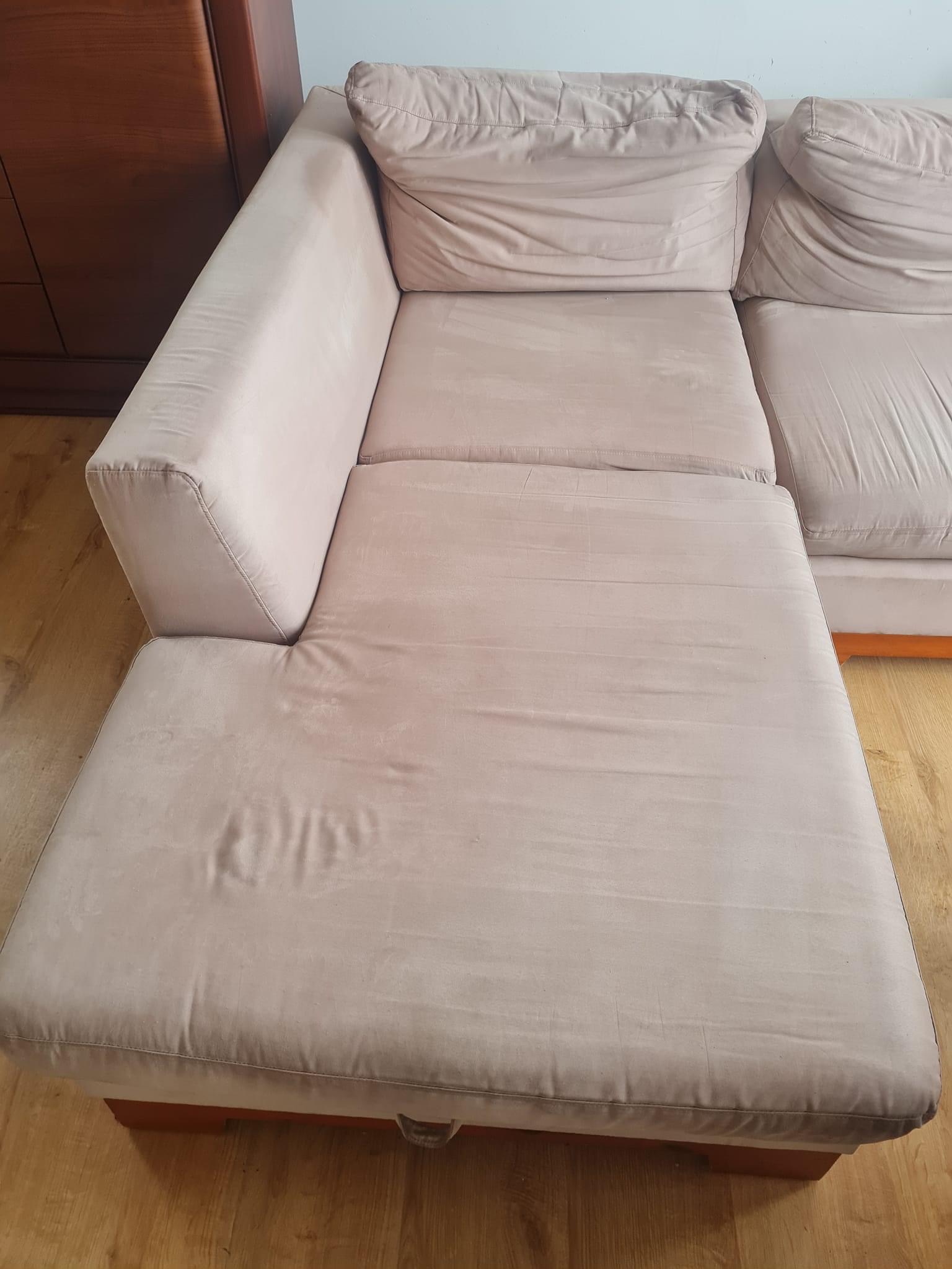 Beżowa, narożna kanapa z widocznymi śladami użytkowania i lekkimi zagnieceniami na siedzisku, stojąca na drewnianej podłodze, z fragmentem drewnianego mebla w tle.
