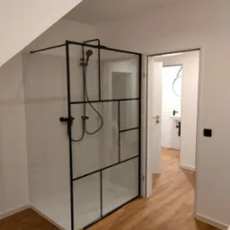 Nowoczesna kabina prysznicowa z czarnymi profilami i armaturą, białe płytki typu metro na ścianach, widok na łazienkę w tle z drewnopodobną podłogą.
