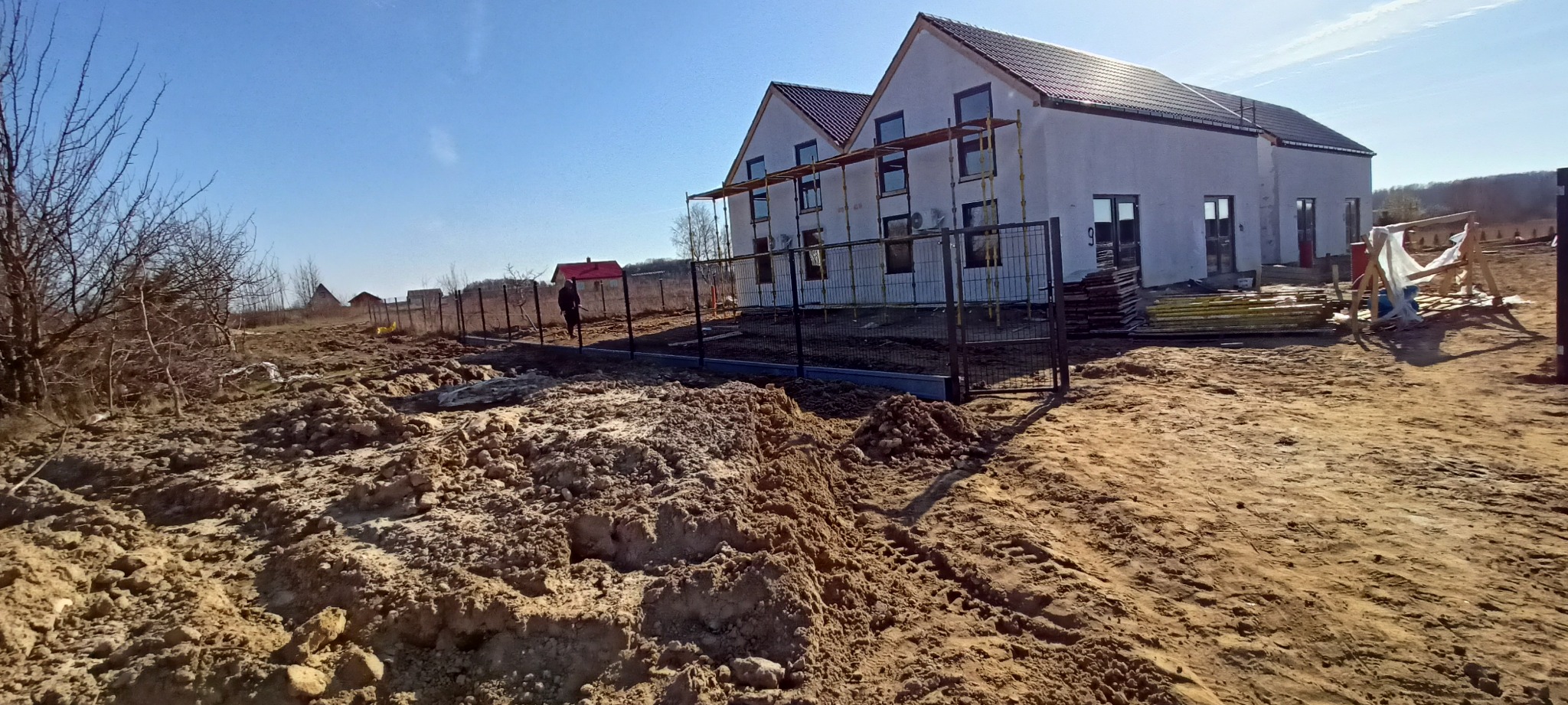 Montaż ogrodzenia panelowego z metalową furtką na nierównym terenie budowy, w tle ocieplony budynek w trakcie prac wykończeniowych.