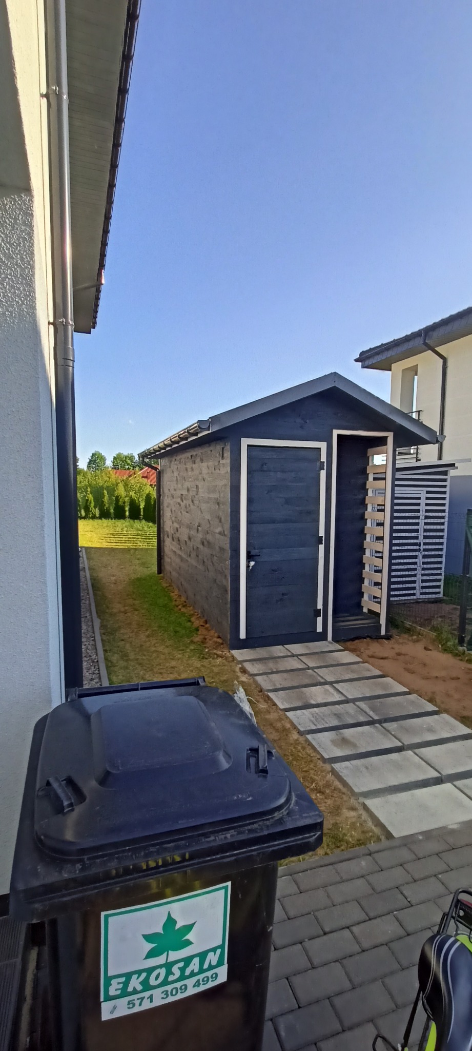 Nowo wybudowana, ciemna, drewniana szopa ogrodowa z białymi obramowaniami drzwi, widziana z perspektywy kosza na śmieci z logo EKOSAN, z ścieżką z betonowych płyt prowadzącą do wejścia.