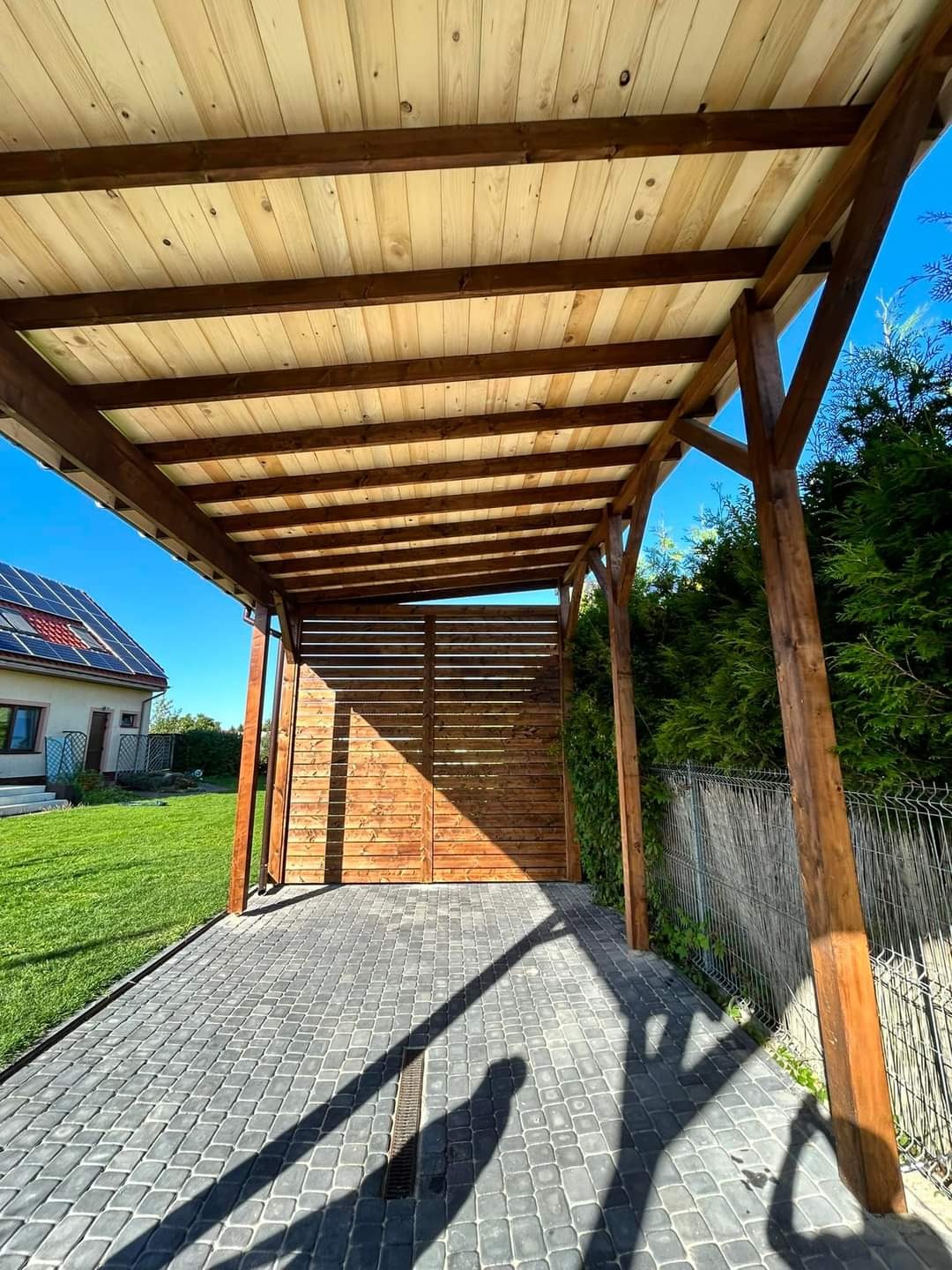 Drewniana wiata garażowa z płaskim dachem i ażurową ścianą, wykończona kostką brukową, w tle dom z panelami słonecznymi i zadbany trawnik.
