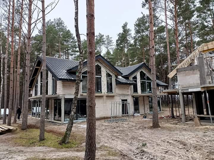 Nowoczesny dom w stanie surowym zamkniętym z ciemną dachówką, otoczony sosnowym lasem, widok z zewnątrz na tle pochmurnego nieba.