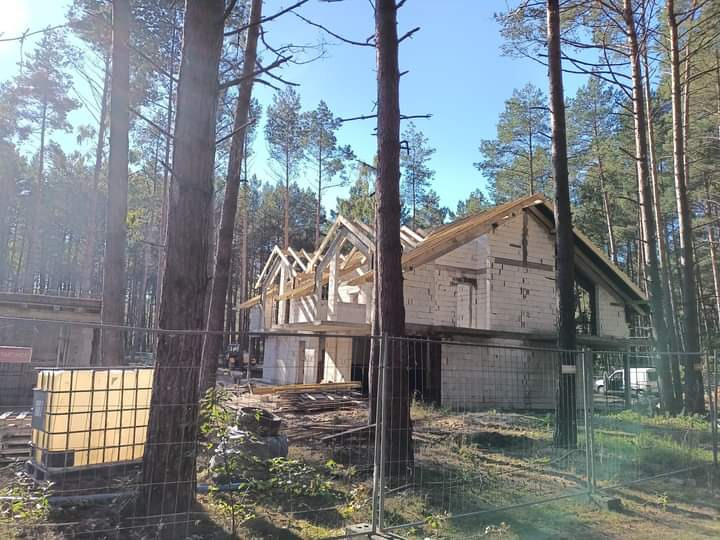 Budowa domu z białych bloczków w lesie sosnowym, widoczne elementy konstrukcji dachu, teren budowy zabezpieczony siatką.