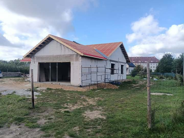 Budynek w trakcie budowy z częściowo położonym dachem z czerwonej dachówki, widoczne rusztowania i konstrukcja więźby dachowej, w tle budynek wielorodzinny.