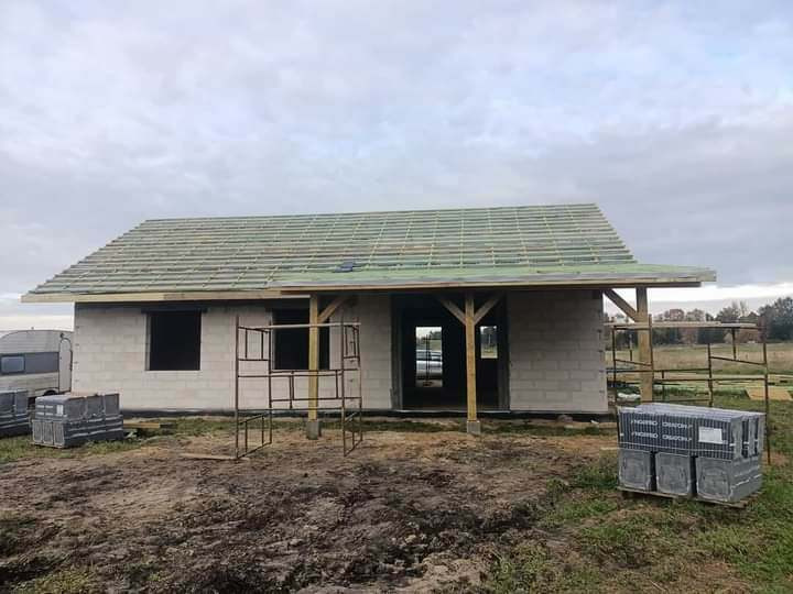 Budowa domu jednorodzinnego w trakcie, widoczne ściany z bloczków, drewniana konstrukcja dachu z łatami, palety z materiałami budowlanymi na trawie, pochmurne niebo.