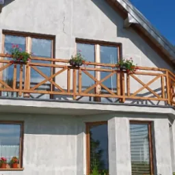 Drewniany balkon z balustradą w kształcie krzyży, ozdobiony doniczkami z czerwonymi kwiatami, na tle surowej elewacji domu.