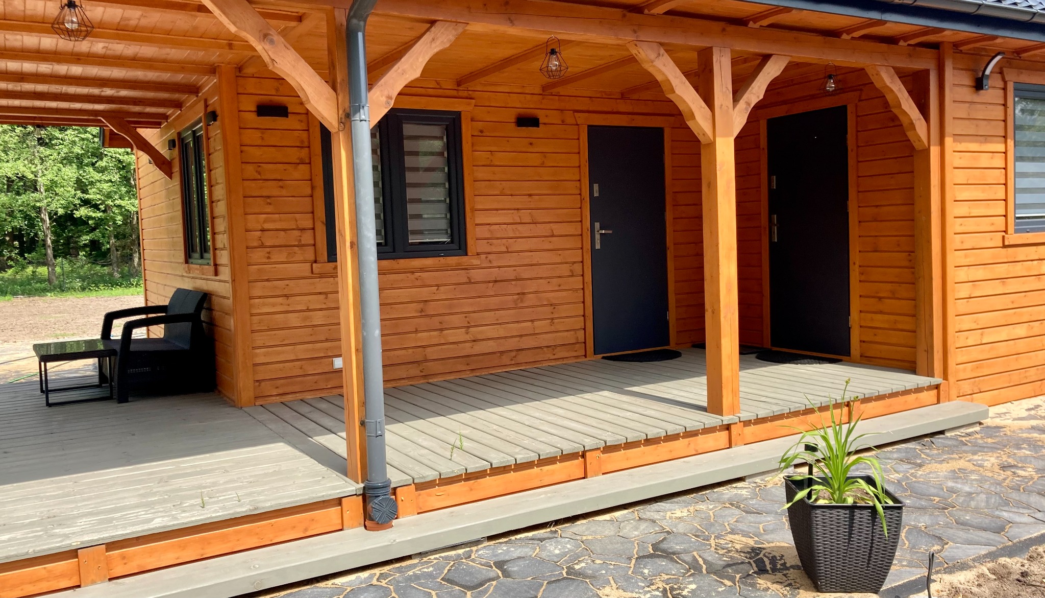 Drewniana konstrukcja z dwoma wejściami, szarą podłogą i czarnymi drzwiami, widoczne elementy oświetlenia i meble ogrodowe, roślina w ozdobnej donicy z przodu.