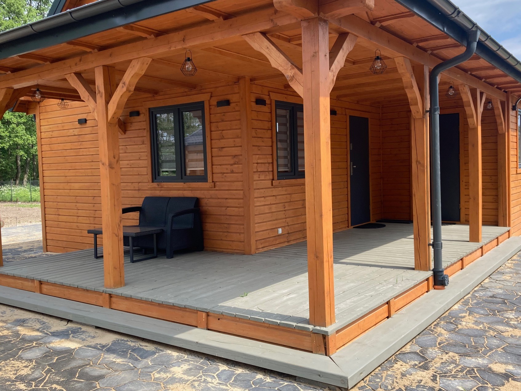 Drewniana konstrukcja z zadaszonym tarasem, wyposażona w czarne meble ogrodowe i oświetlenie w stylu industrialnym, wykończona w ciepłym odcieniu drewna, z widocznym fragmentem kamiennej ścieżki.