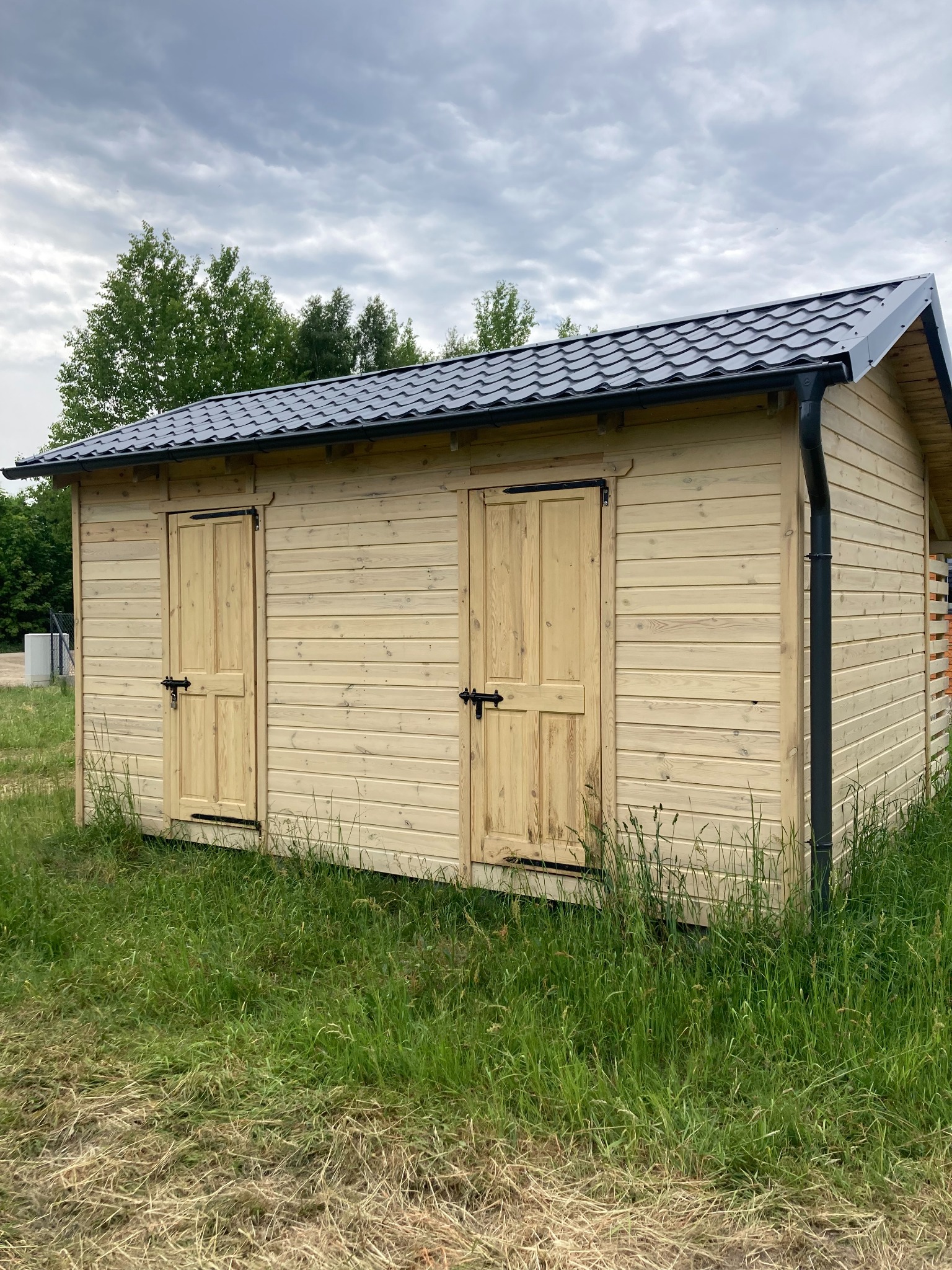 Nowa, drewniana altana z dwoma wejściami, pokryta blachodachówką w kolorze grafitowym, z widocznymi rynnami, otoczona wysoką trawą.