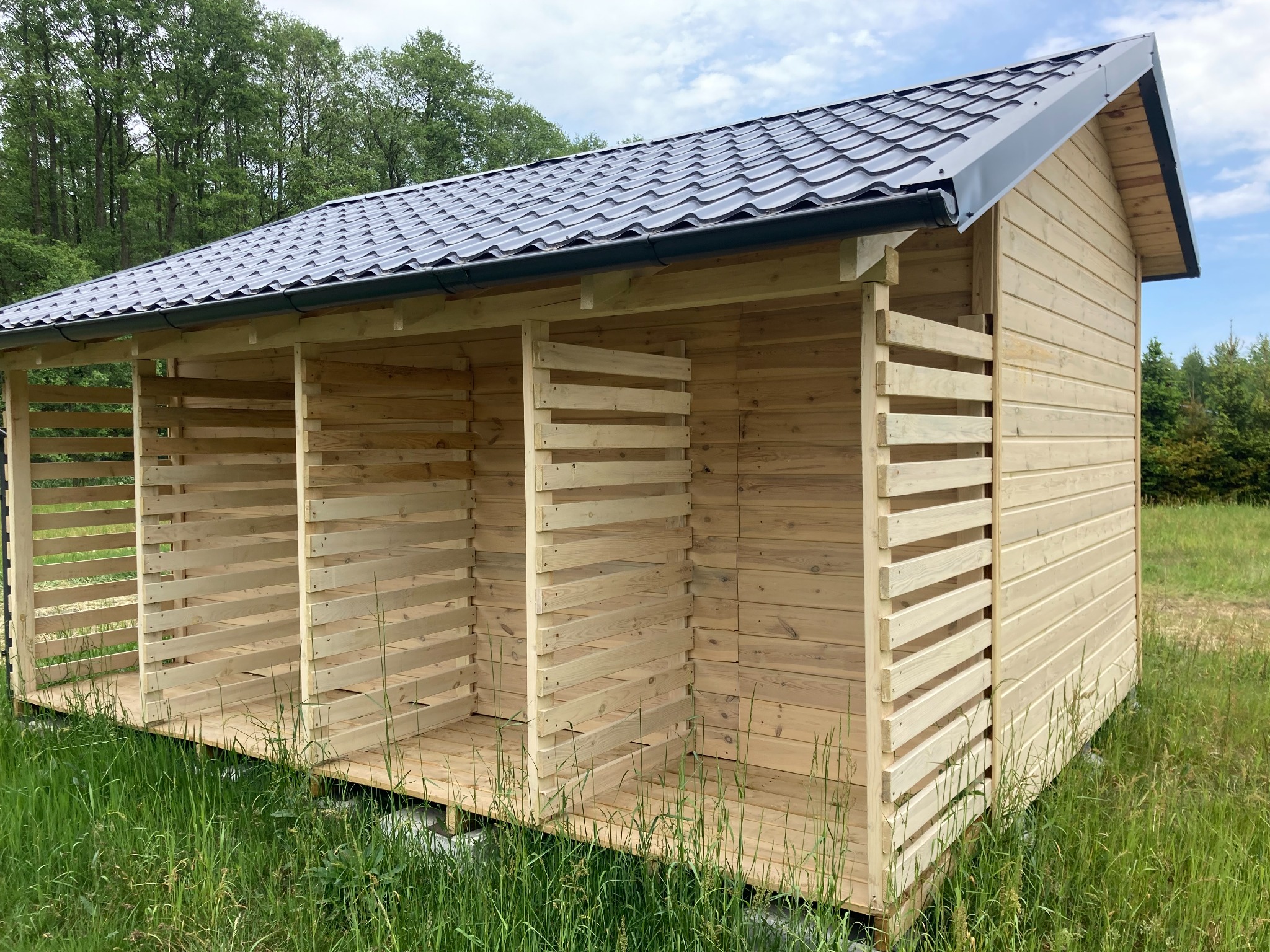 Surowa drewniana konstrukcja z trzema otwartymi przegrodami i metalowym dachem, prawdopodobnie przeznaczona do przechowywania drewna, otoczona zieloną trawą.