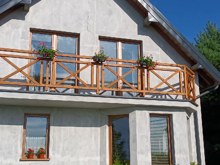 Drewniany balkon z balustradą w kształcie krzyży, ozdobiony doniczkami z czerwonymi kwiatami, na tle surowej elewacji domu.