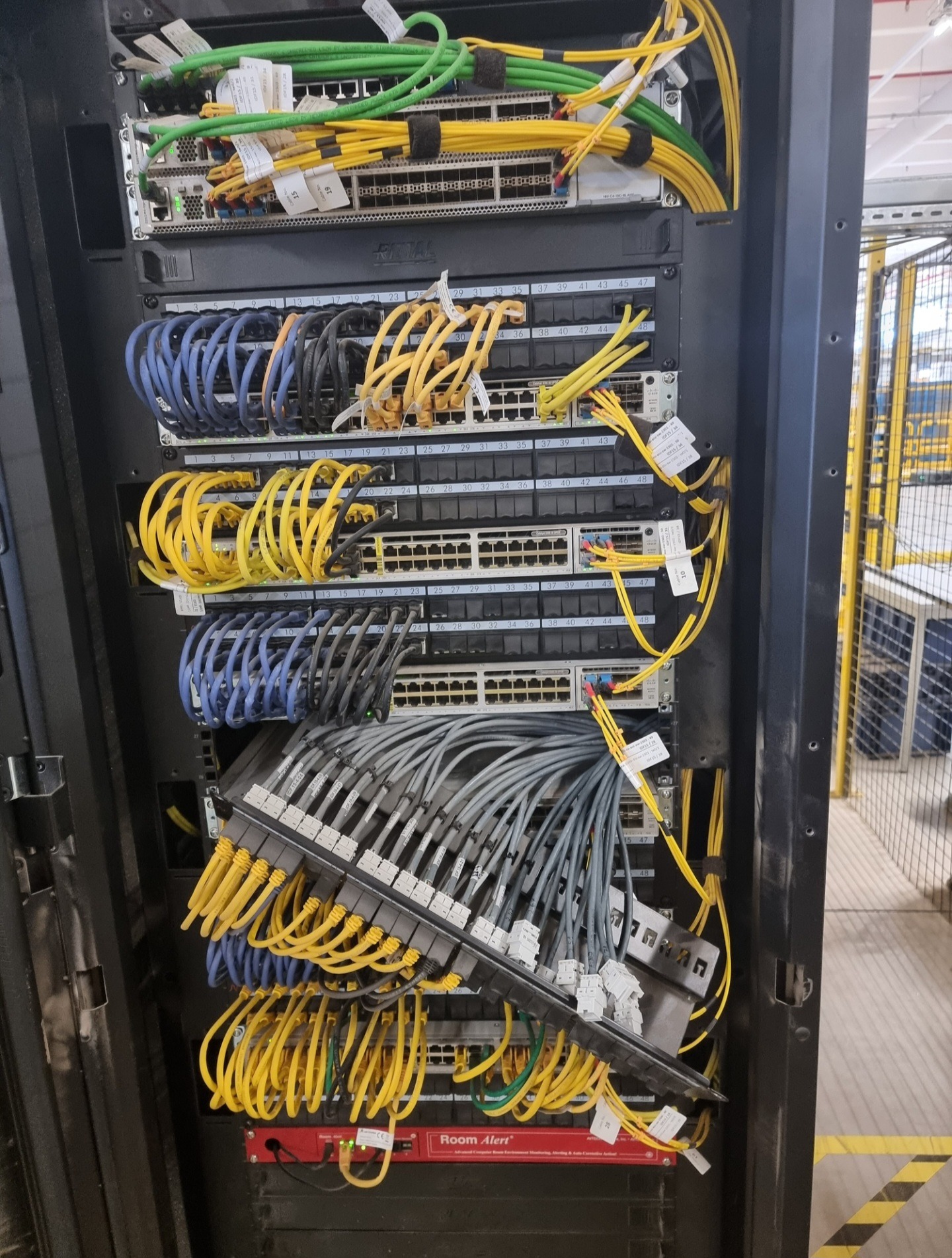 Szafa rack z zamontowanymi przełącznikami sieciowymi i okablowaniem strukturalnym w kolorach żółtym, niebieskim i zielonym, z widocznym panelem krosowniczym i modułem Room Alert na dole.