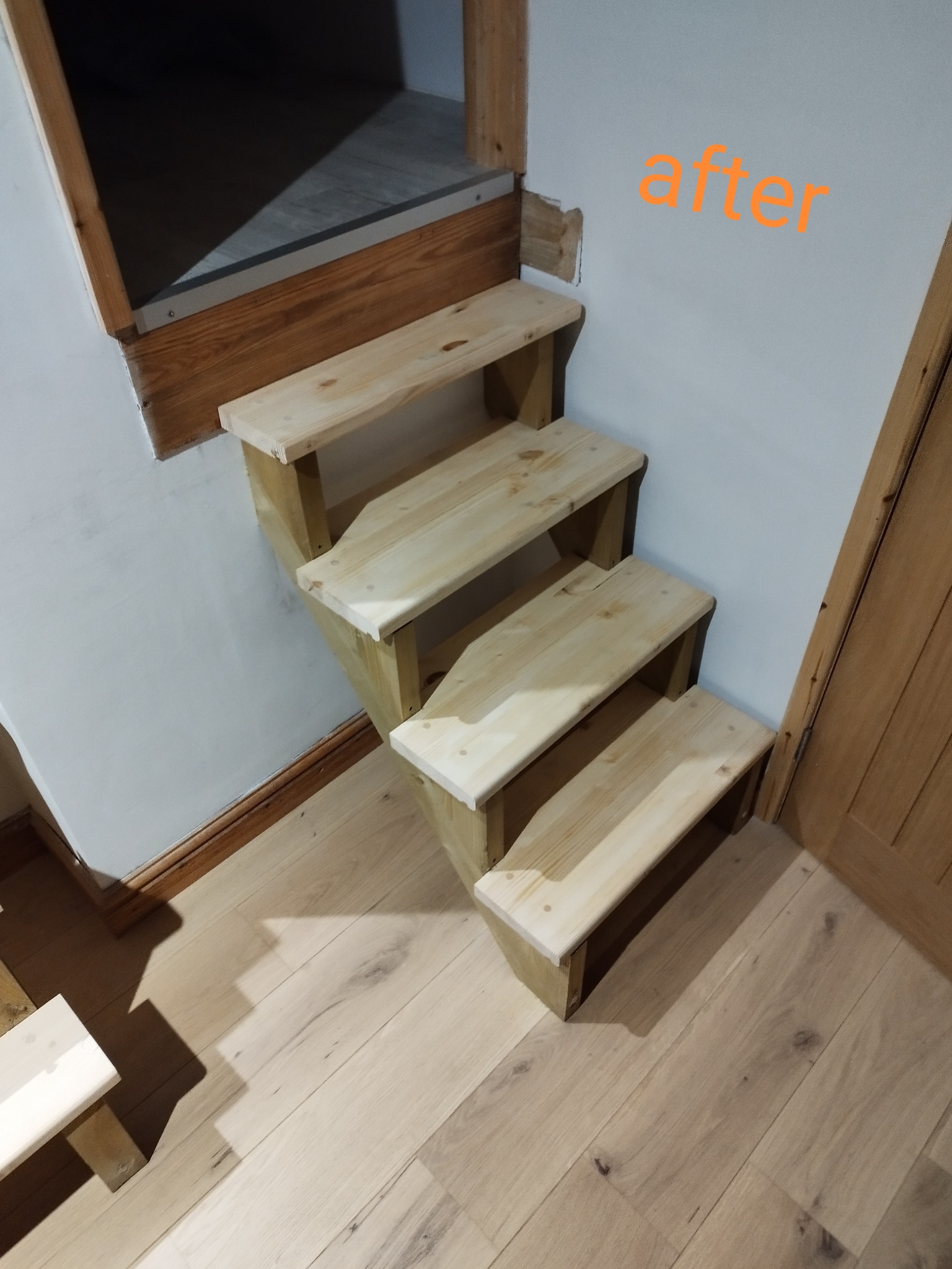 Drewniane schody z surowego drewna prowadzące do niskiego otworu, widoczne ślady naprawy ściany, podpis 'after' na ścianie.