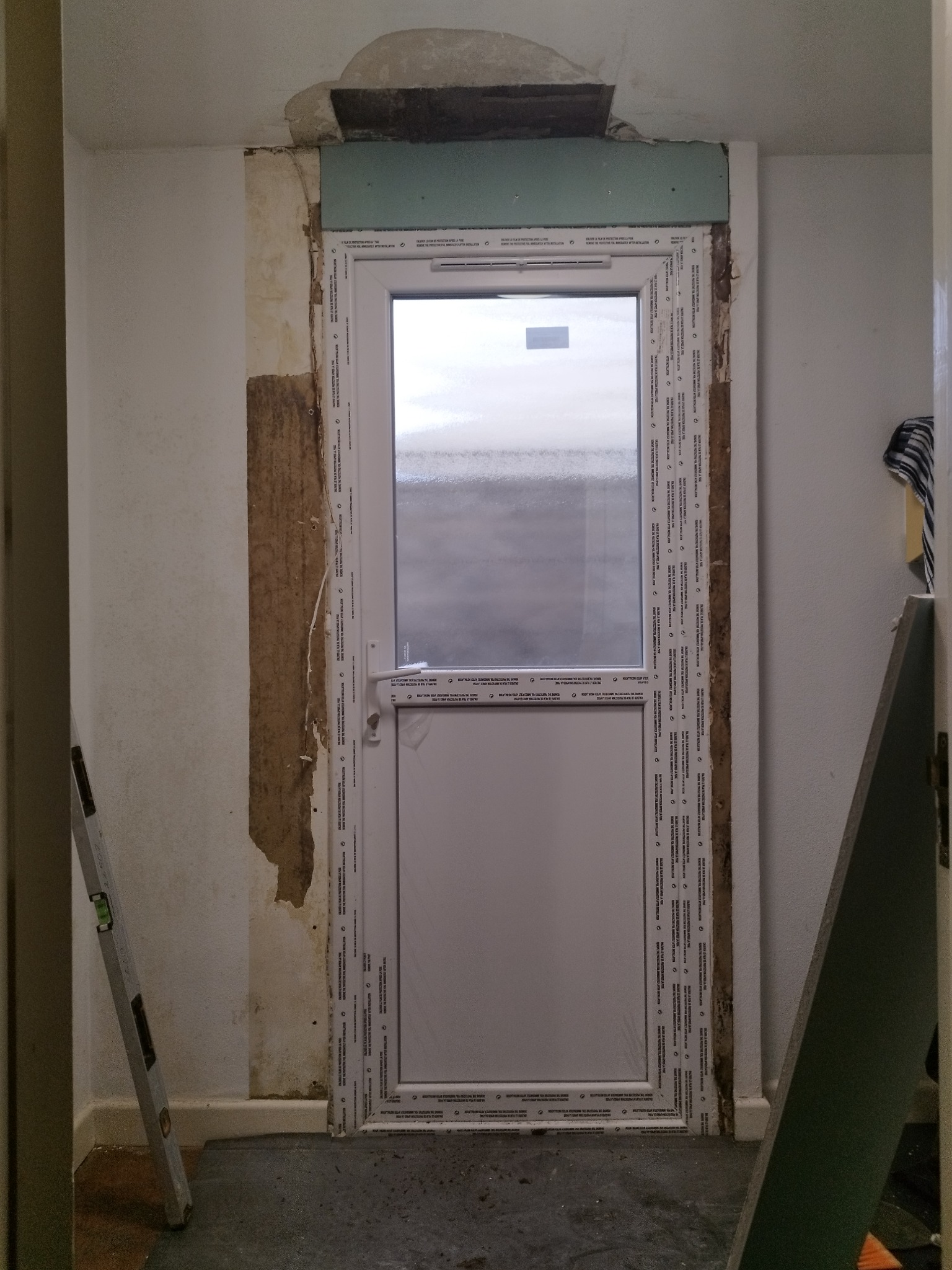 Montaż nowych drzwi balkonowych z plastikową ramą w trakcie remontu. Widoczne uszkodzenia ściany i sufitu, poziomica oparta o ścianę.