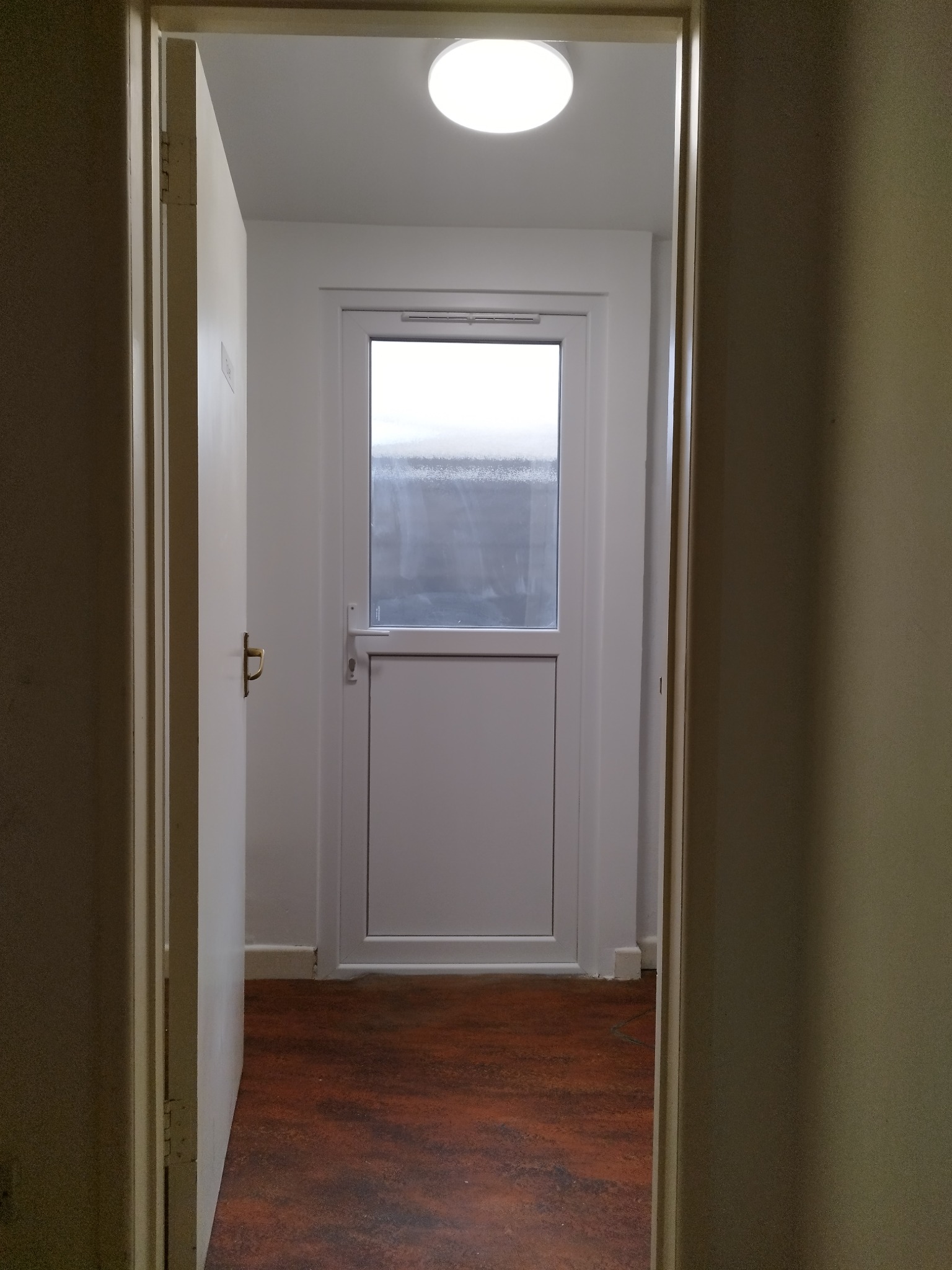 Wnętrze korytarza z widokiem na białe drzwi z szybą i okrągłą lampą sufitową, widziane zza uchylonych drzwi po lewej stronie.