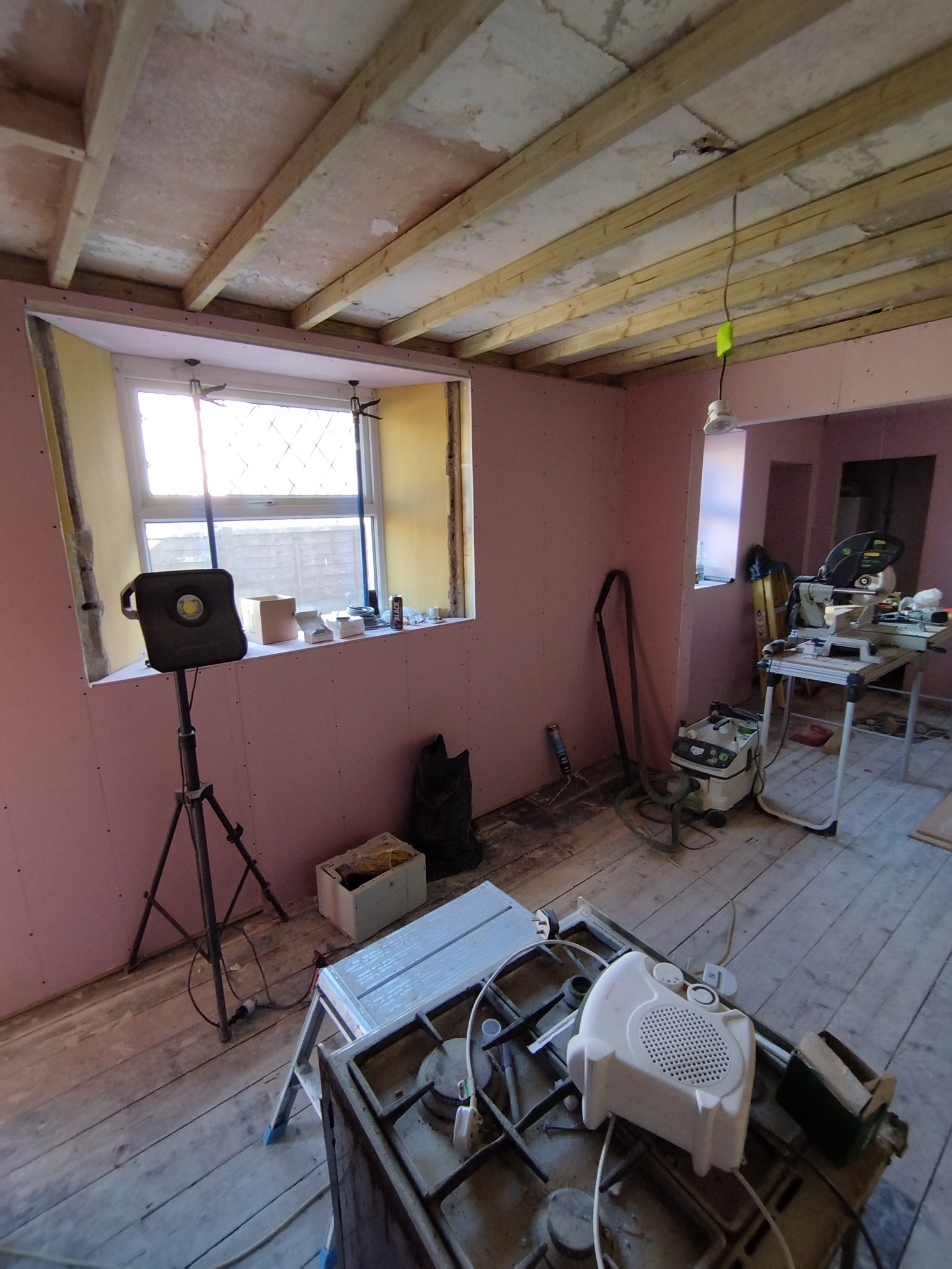 Wnętrze pokoju w trakcie remontu, ściany wyłożone płytami gipsowo-kartonowymi w kolorze różowym, surowy drewniany sufit, okno z kratką, widoczne narzędzia i sprzęt budowlany, w tym lampa...