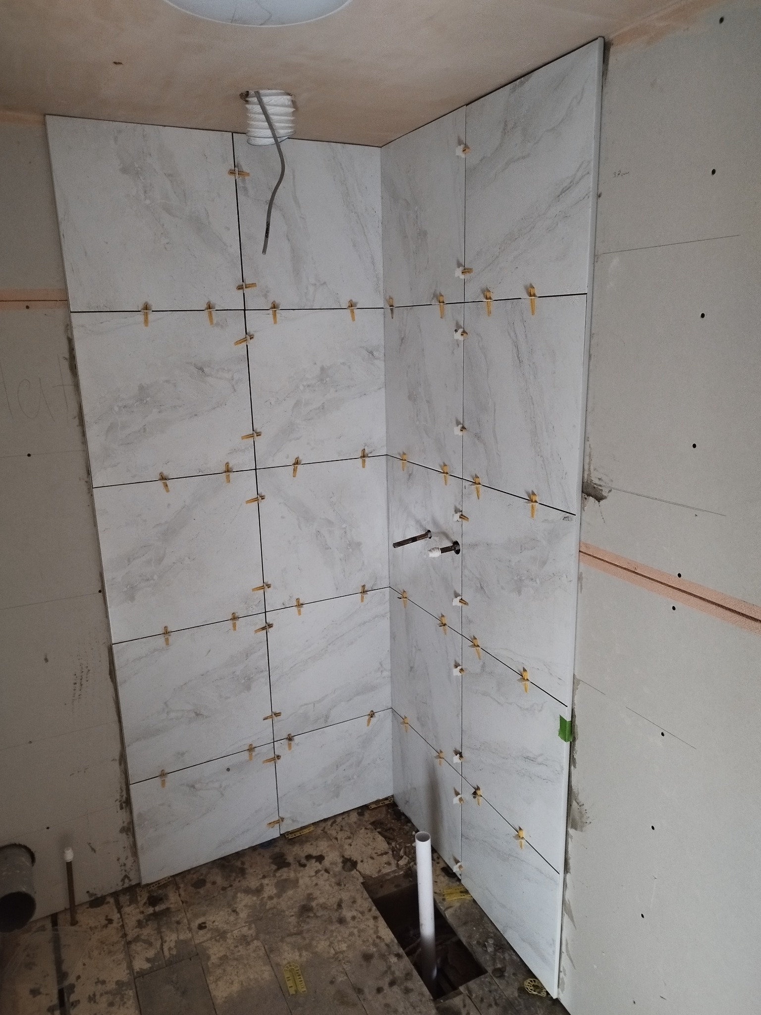 Ściana łazienki w trakcie wykańczania, z zamontowanymi płytkami imitującymi marmur, z krzyżykami dystansowymi, widoczne instalacje wodne i fragment surowej podłogi.