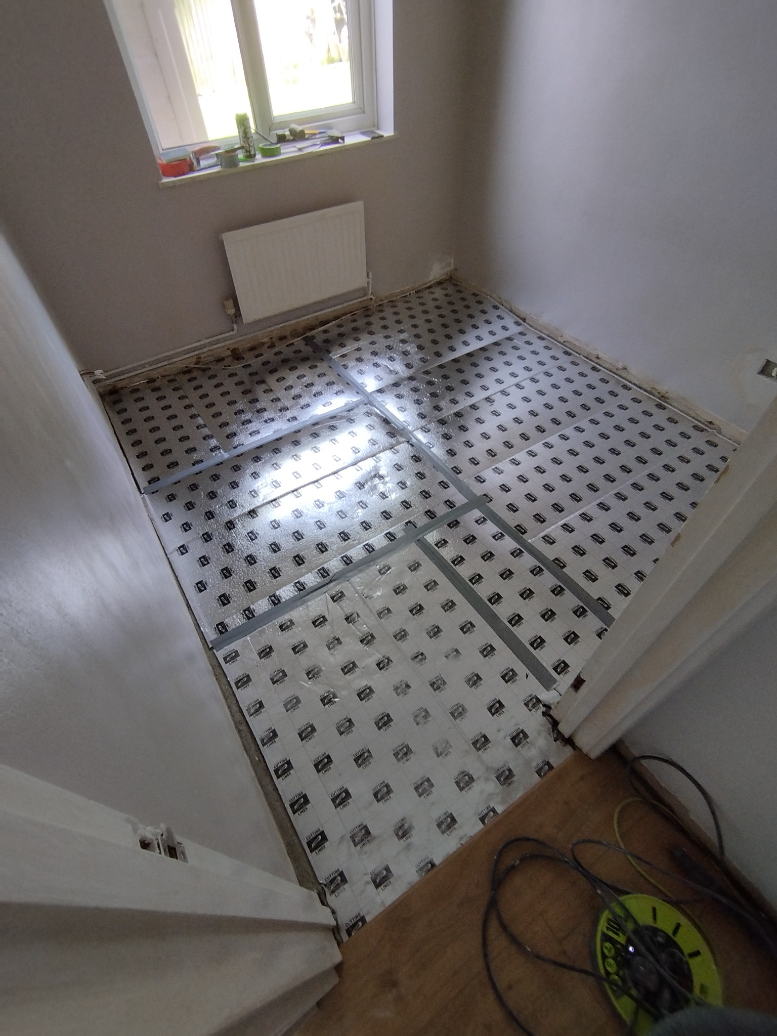 Przygotowanie podłoża pod panele: rozłożona folia izolacyjna z nadrukiem i listwy dystansowe w małym pokoju z oknem i grzejnikiem.