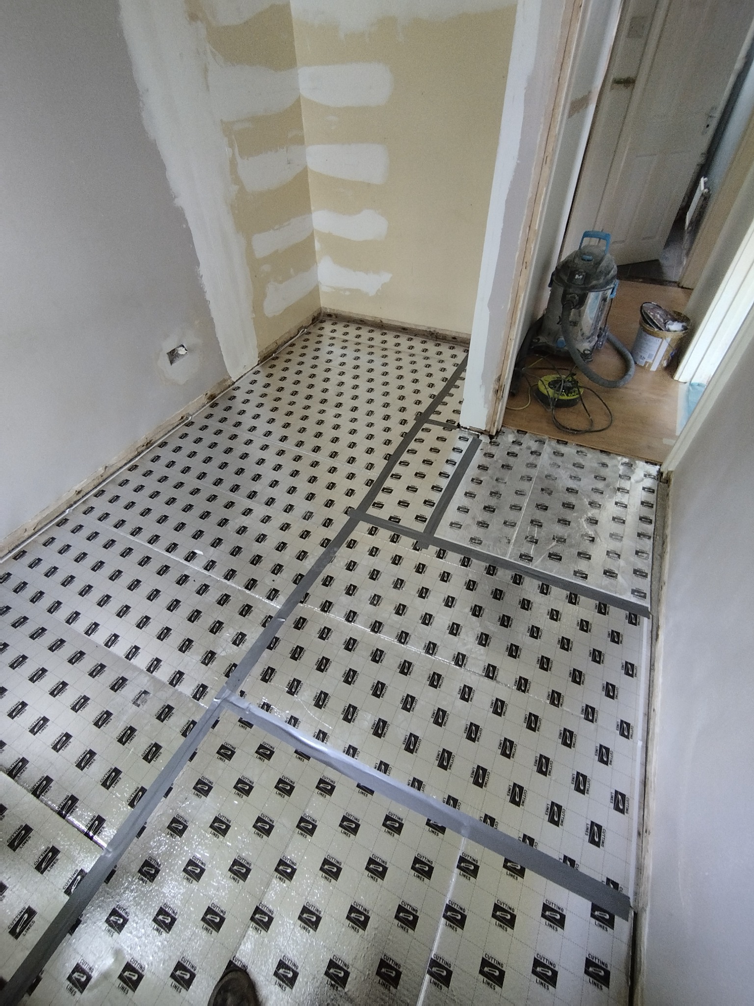 Przygotowanie podłoża pod panele: rozłożona folia izolacyjna z nadrukiem i taśmy wyznaczające obszary, ściany w trakcie szpachlowania, widoczny odkurzacz przemysłowy.