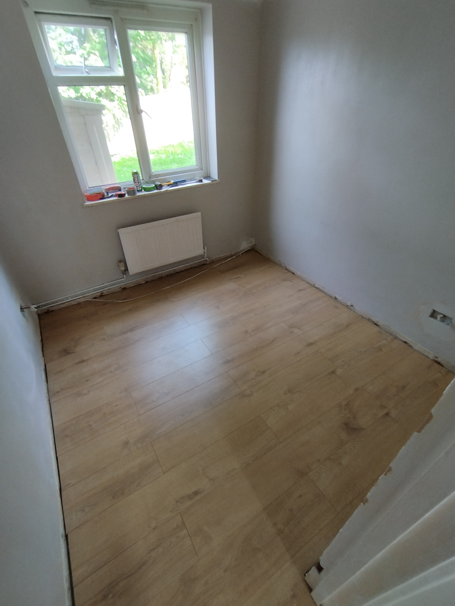 Pokój po ułożeniu paneli podłogowych w odcieniach jasnego drewna, widoczne niedoskonałości przy ścianach i listwach przypodłogowych, okno z widokiem na zieleń za nim.