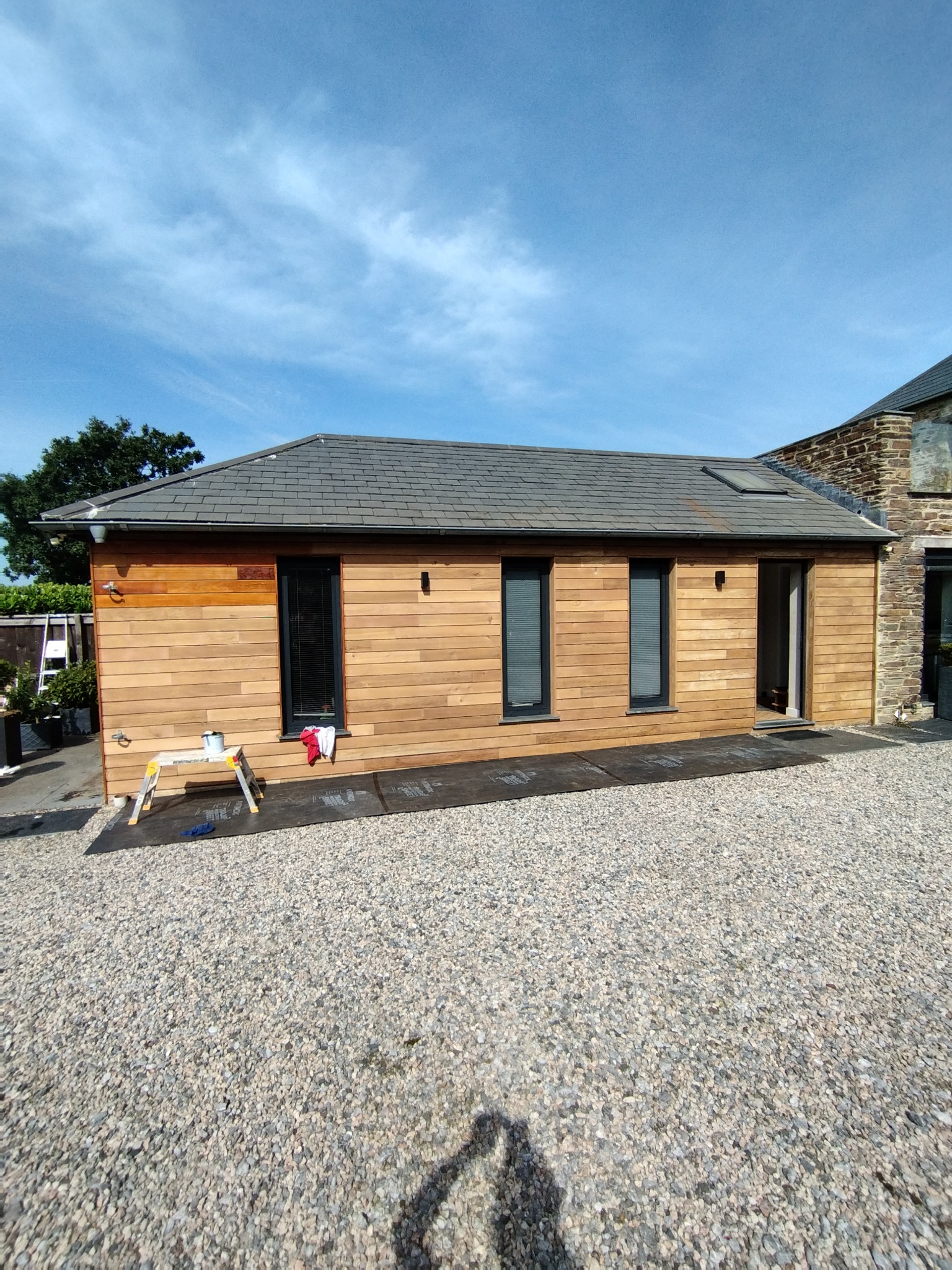 Nowoczesny budynek z drewnianą elewacją w trakcie impregnacji, widoczny fragment elewacji z surowego drewna i fragment po impregnacji, na pierwszym planie rozłożony stołek malarski z puszką...