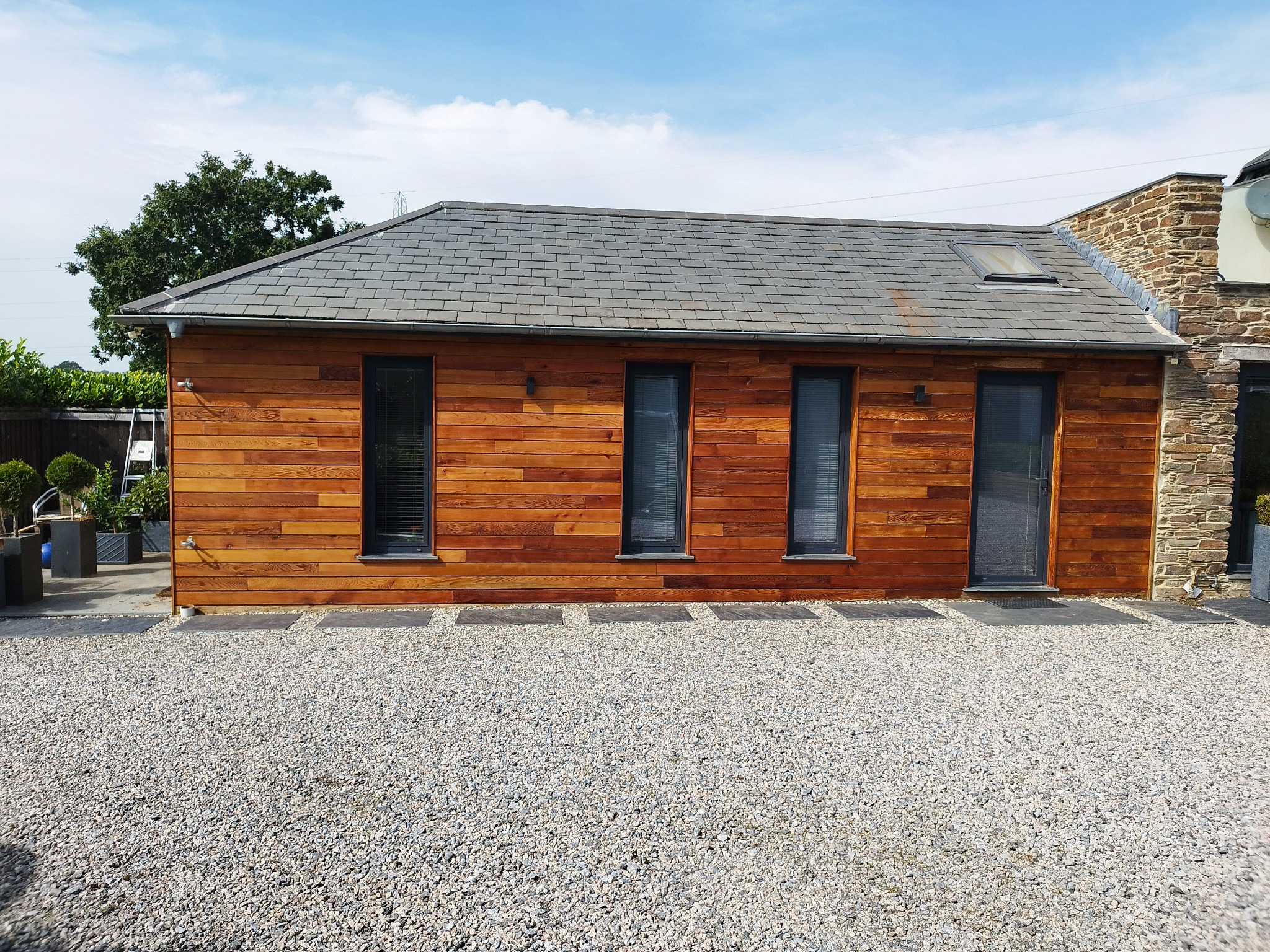 Nowoczesny budynek z drewnianą elewacją i szarym dachem, czarne okna z żaluzjami, kamienny fragment ściany, podłoże wysypane żwirem.