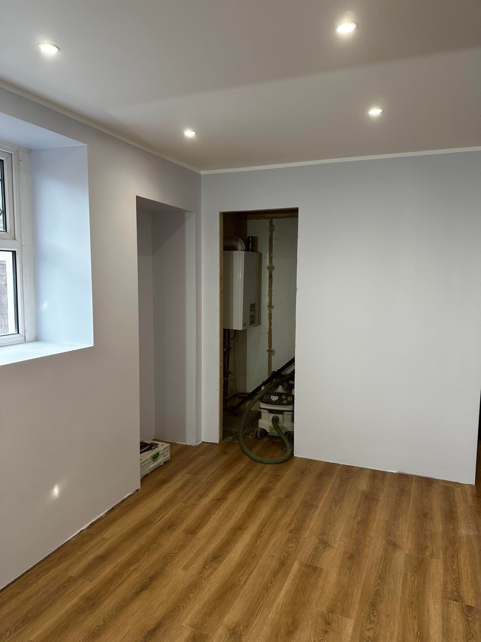 Wnętrze pokoju po remoncie: świeżo pomalowane szare ściany, ułożone panele podłogowe, widoczny fragment okna i otwór drzwiowy z kotłem gazowym w tle oraz odkurzacz przemysłowy.