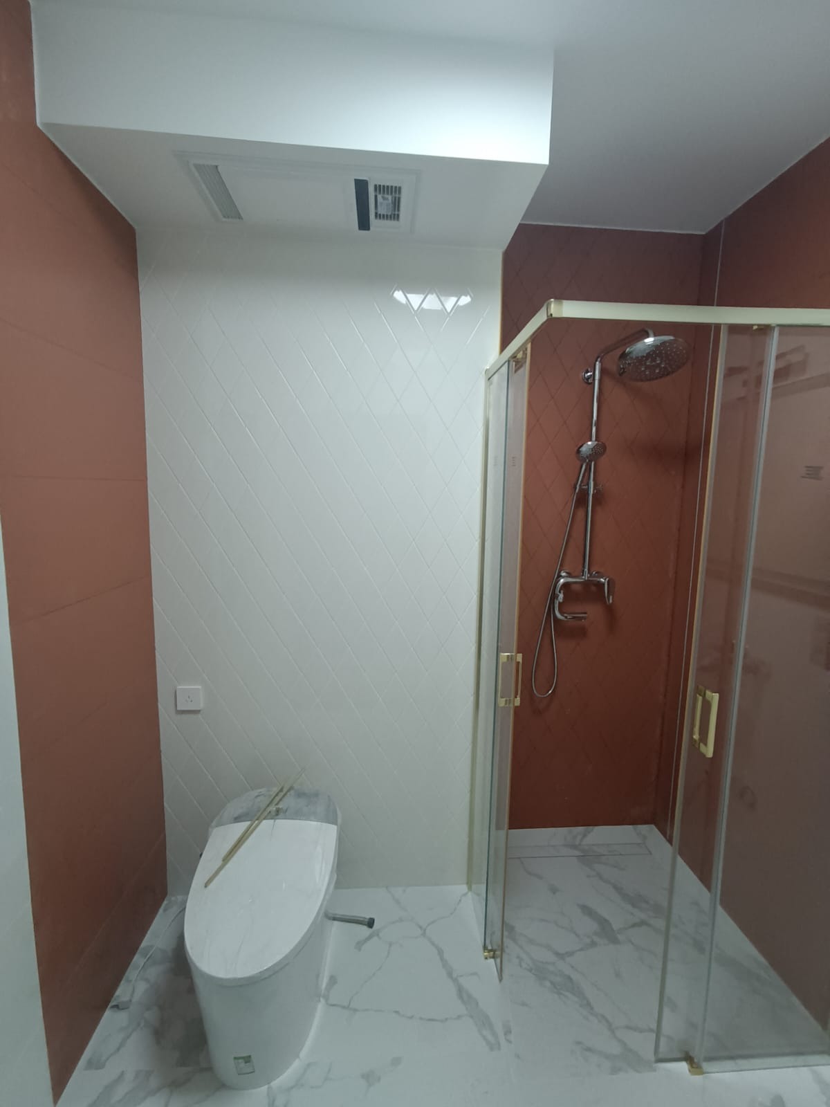 Nowoczesna łazienka po remoncie z biało-brązowymi płytkami, kabiną prysznicową ze złotymi uchwytami i podwieszaną toaletą z deską sedesową zabezpieczoną folią.