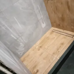 Wykończona kabina prysznicowa z szarymi płytkami imitującymi marmur na ścianach, drewnopodobnymi płytkami na podłodze i ścianie oraz czarnym odpływem liniowym.