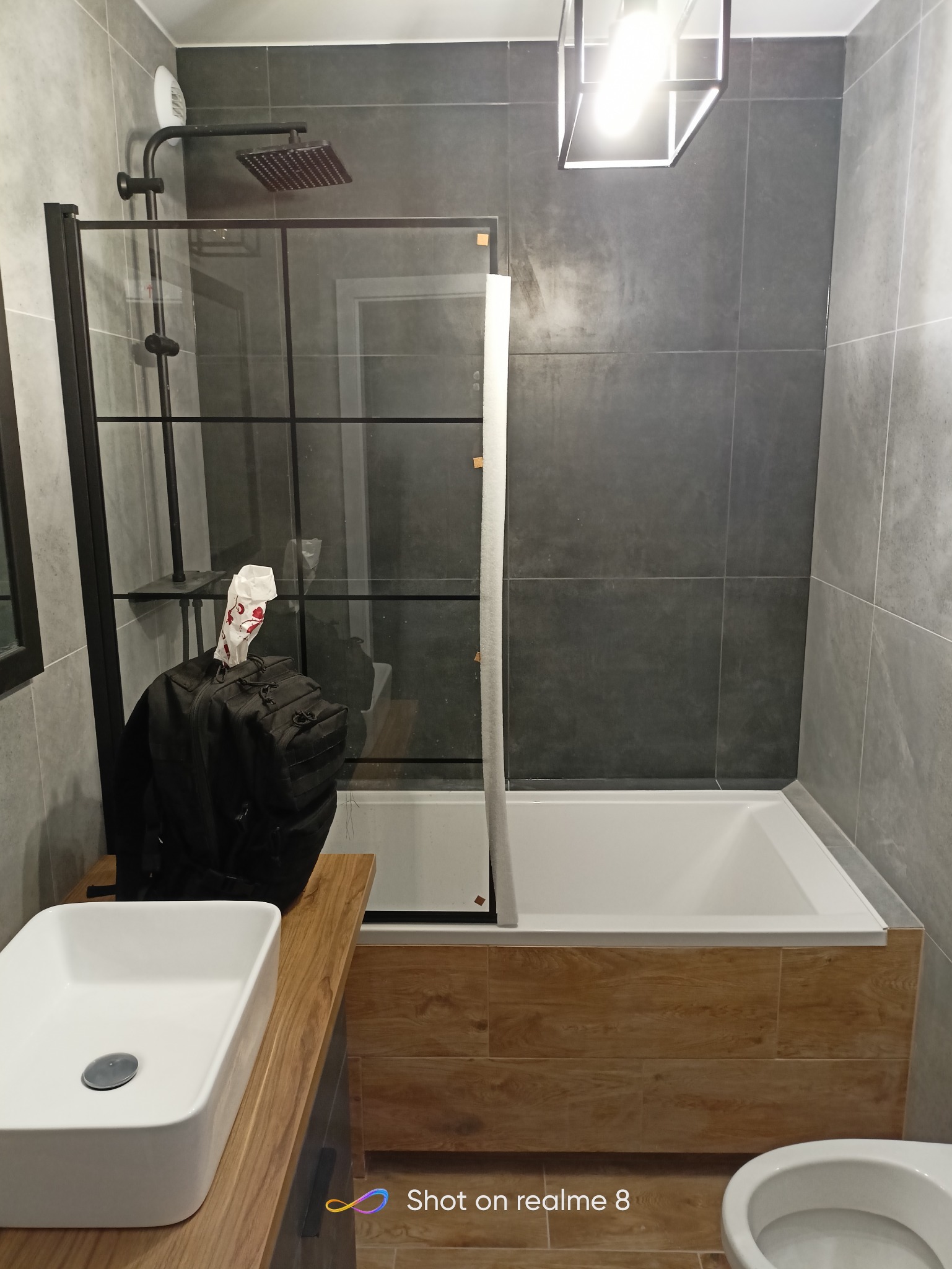 Nowoczesna łazienka z szarą glazurą, kabiną prysznicową typu walk-in z czarnymi profilami, umywalką nablatową na drewnianym blacie i wiszącą lampą. Na blacie leży czarny plecak.