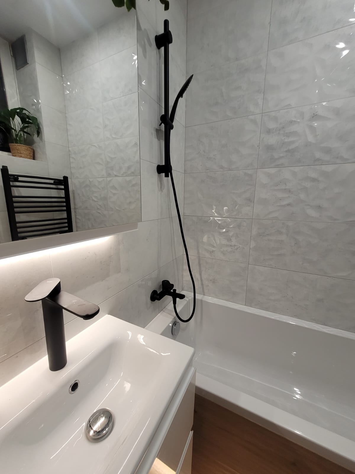 Nowoczesna łazienka z białymi płytkami o trójwymiarowej fakturze, czarną armaturą, umywalką nablatową i grzejnikiem drabinkowym odbijającym się w lustrze.