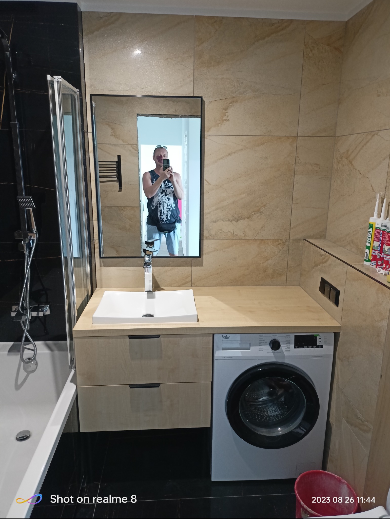 Ukończona łazienka z beżowymi płytkami, czarną armaturą prysznicową, umywalką nablatową, szafką z jasnego drewna i pralką Beko obok, widoczna osoba robiąca zdjęcie w lustrze.