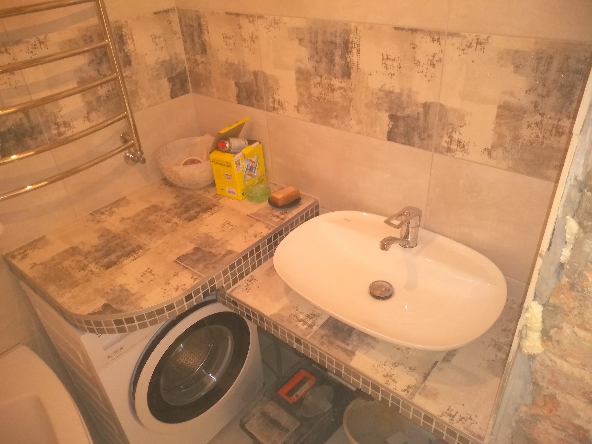 Umywalka nablatowa z baterią, obok pralka z blatem wykończonym mozaiką, na ścianie płytki imitujące kamień, w tle grzejnik drabinkowy i chemia gospodarcza.