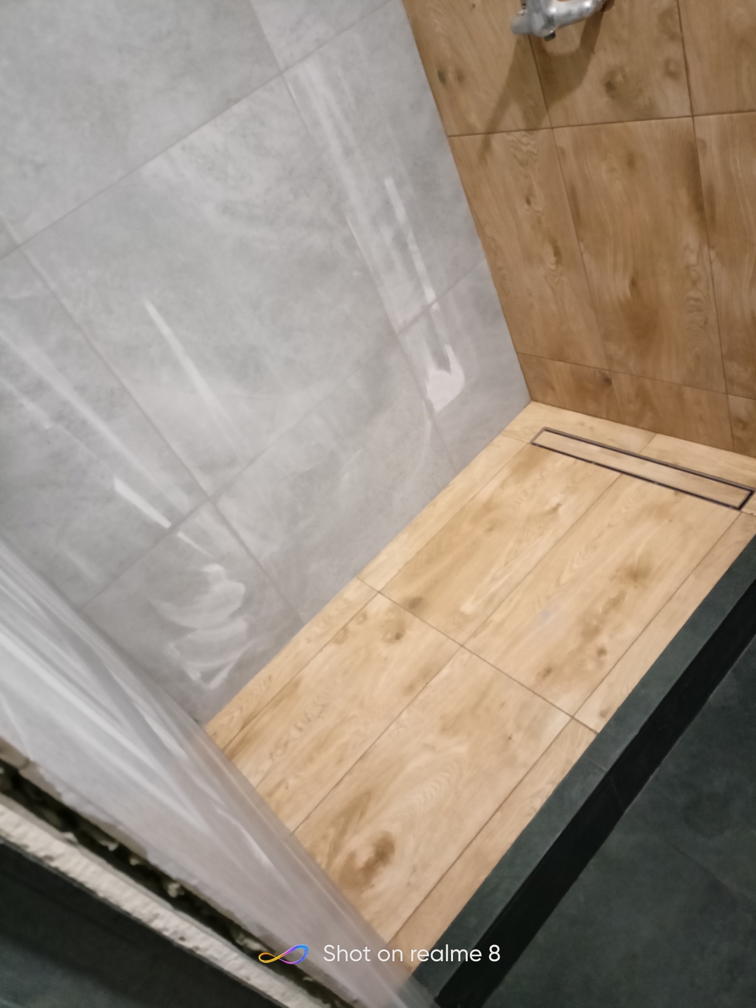 Wykończona kabina prysznicowa z szarymi płytkami imitującymi marmur na ścianach, drewnopodobnymi płytkami na podłodze i ścianie oraz czarnym odpływem liniowym.