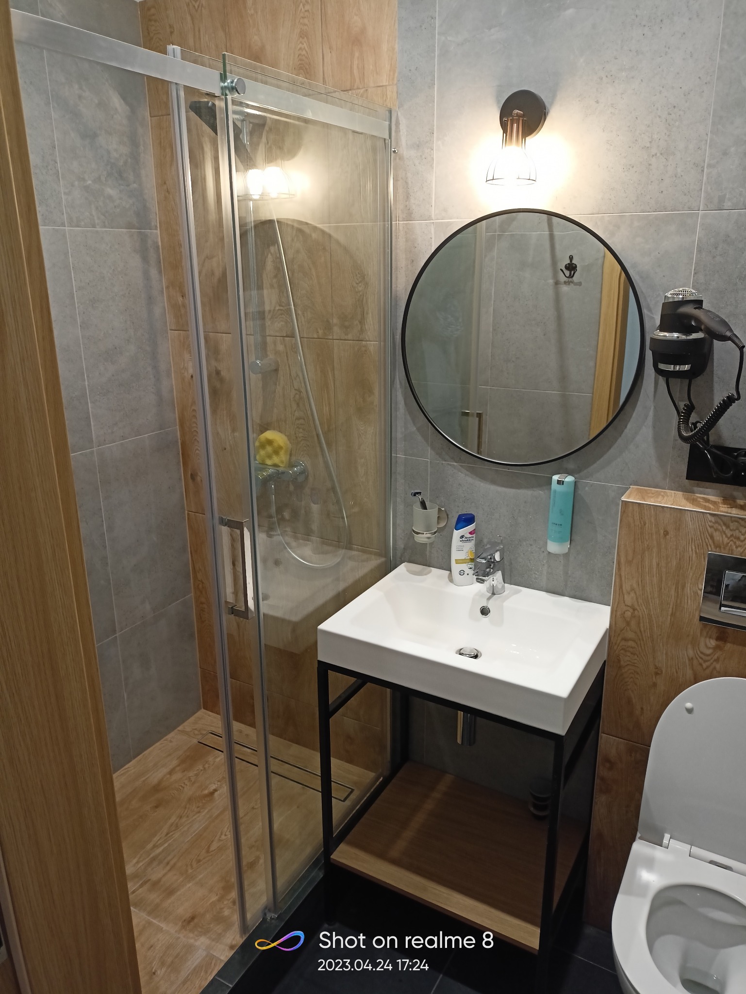 Nowoczesna łazienka z kabiną prysznicową typu walk-in, białą umywalką na czarnym stelażu, okrągłym lustrem i szarymi płytkami imitującymi drewno na ścianach.