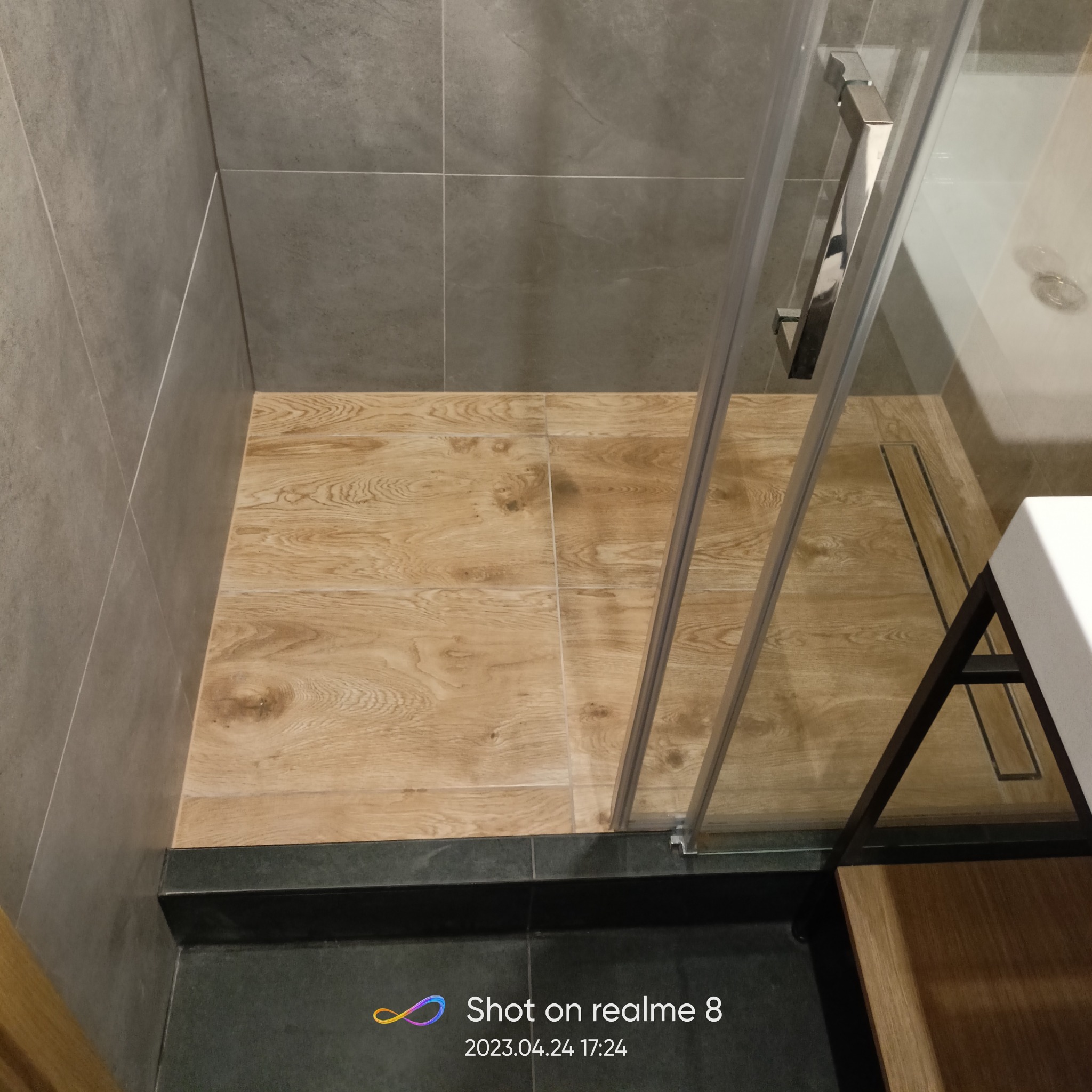 Kabina prysznicowa z drewnopodobną podłogą i szarymi płytkami ściennymi, widoczny odpływ liniowy i fragment szafki umywalkowej.