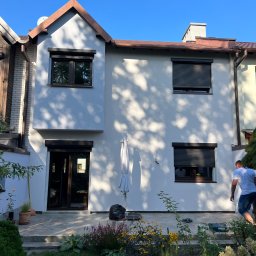 Kamil Jura - Dwukondygnacyjny dom z jasną elewacją i brązowymi elementami wykończeniowymi, widoczne zamknięte rolety okienne, ogród z kwiatami na pierwszym planie, mężczyzna w tle.