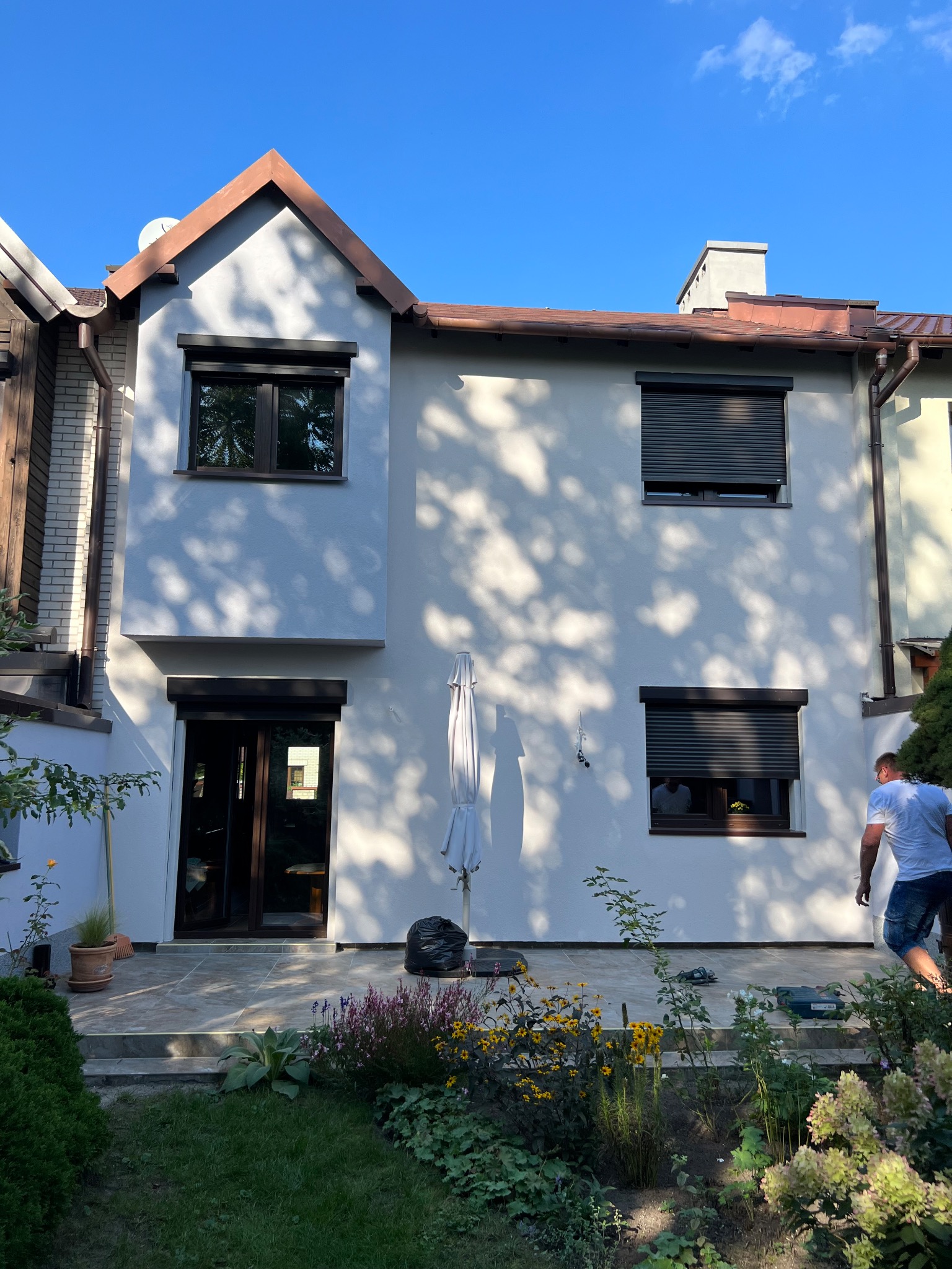Dwukondygnacyjny dom z jasną elewacją i brązowymi elementami wykończeniowymi, widoczne zamknięte rolety okienne, ogród z kwiatami na pierwszym planie, mężczyzna w tle.