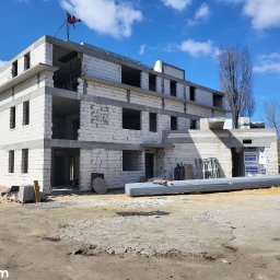 fast.bud.telehiibohdan - Wznoszony budynek z białych bloczków, widoczne otwory okienne i drzwiowe, na placu budowy materiały budowlane i żuraw. Ujęcie w słoneczny dzień.