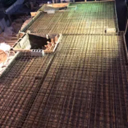 Zbrojenie płyty fundamentowej z widocznym otworem na instalacje, oświetlone sztucznym światłem w nocy, z fragmentami szalunku i bloczków betonowych.